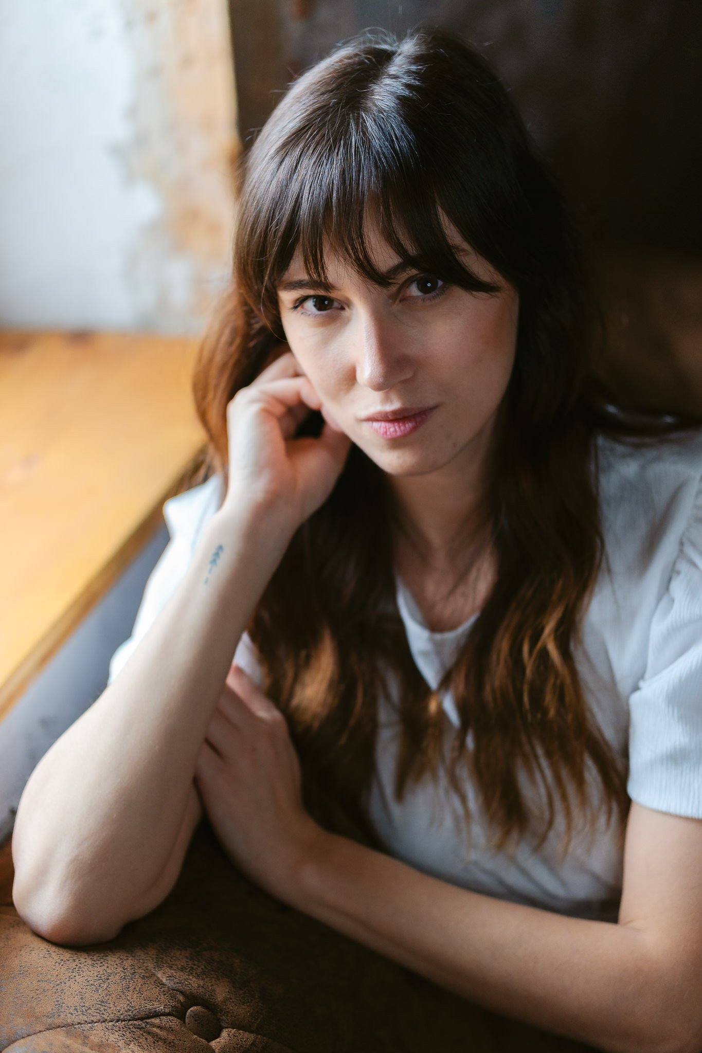 Porträt einer Frau mit braunen Haaren und weißen T-Shirt, die auf einem Sofa sitzt, mit Blick in die Kamera, in einem Raum mit rustikaler Wand.