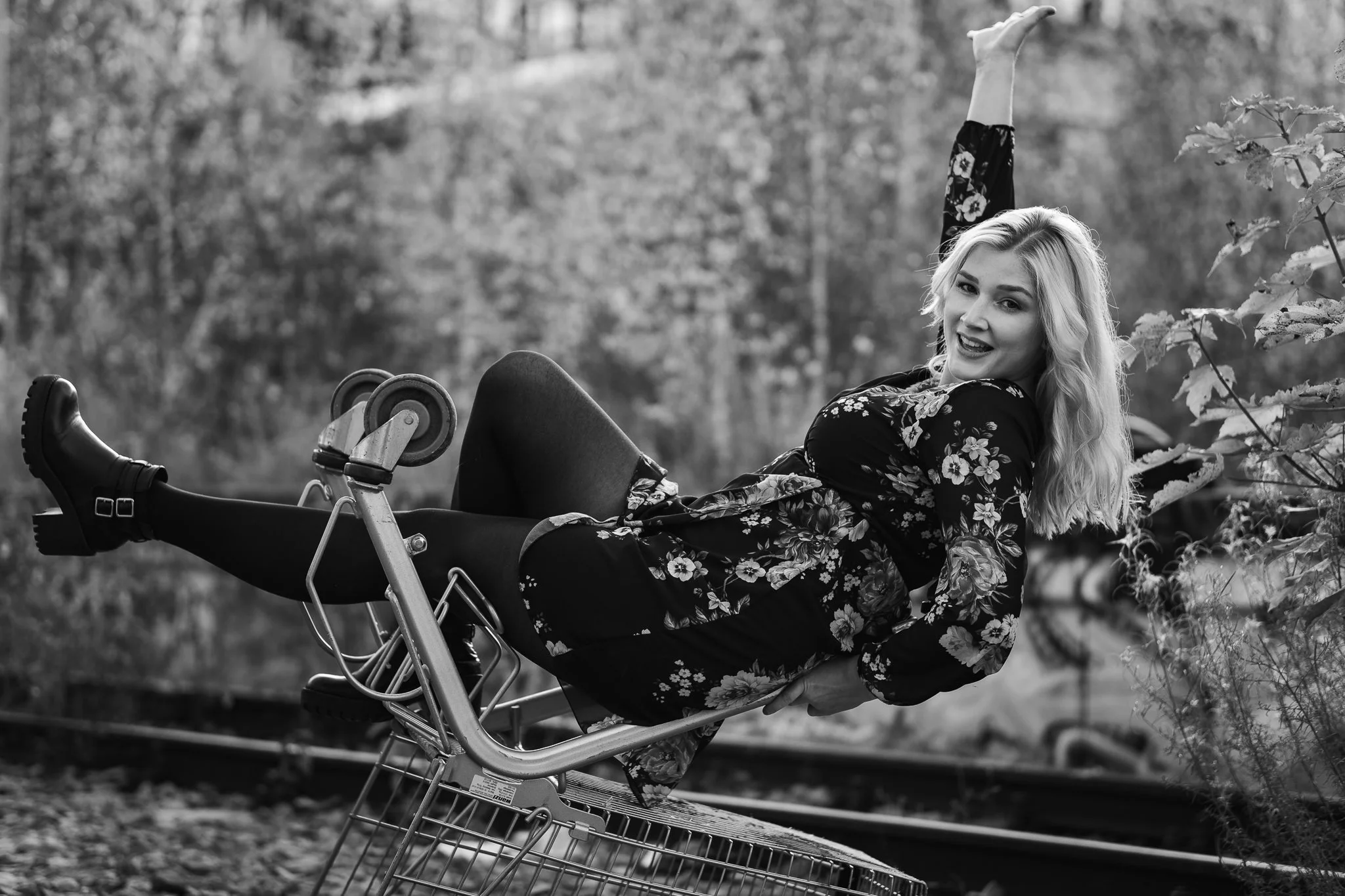 Schwarz-weiß-Foto einer lachenden Frau in einem floral bedruckten Kleid, die auf einem Einkaufswagen in der Natur steht, neben Bahngleisen.