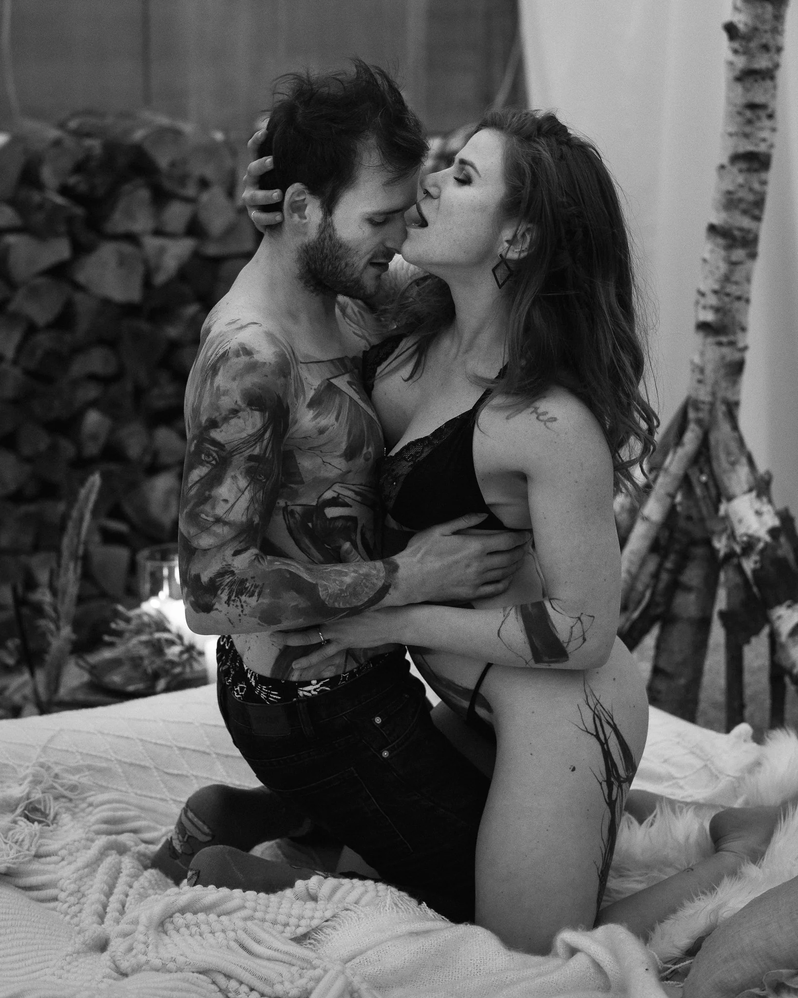 Ein Mann und eine Frau liegen sich nackt auf einem Bett, umarmt und küssend, mit Tattoos auf dem Körper, im Hintergrund ein Kamin mit Holzscheiten.