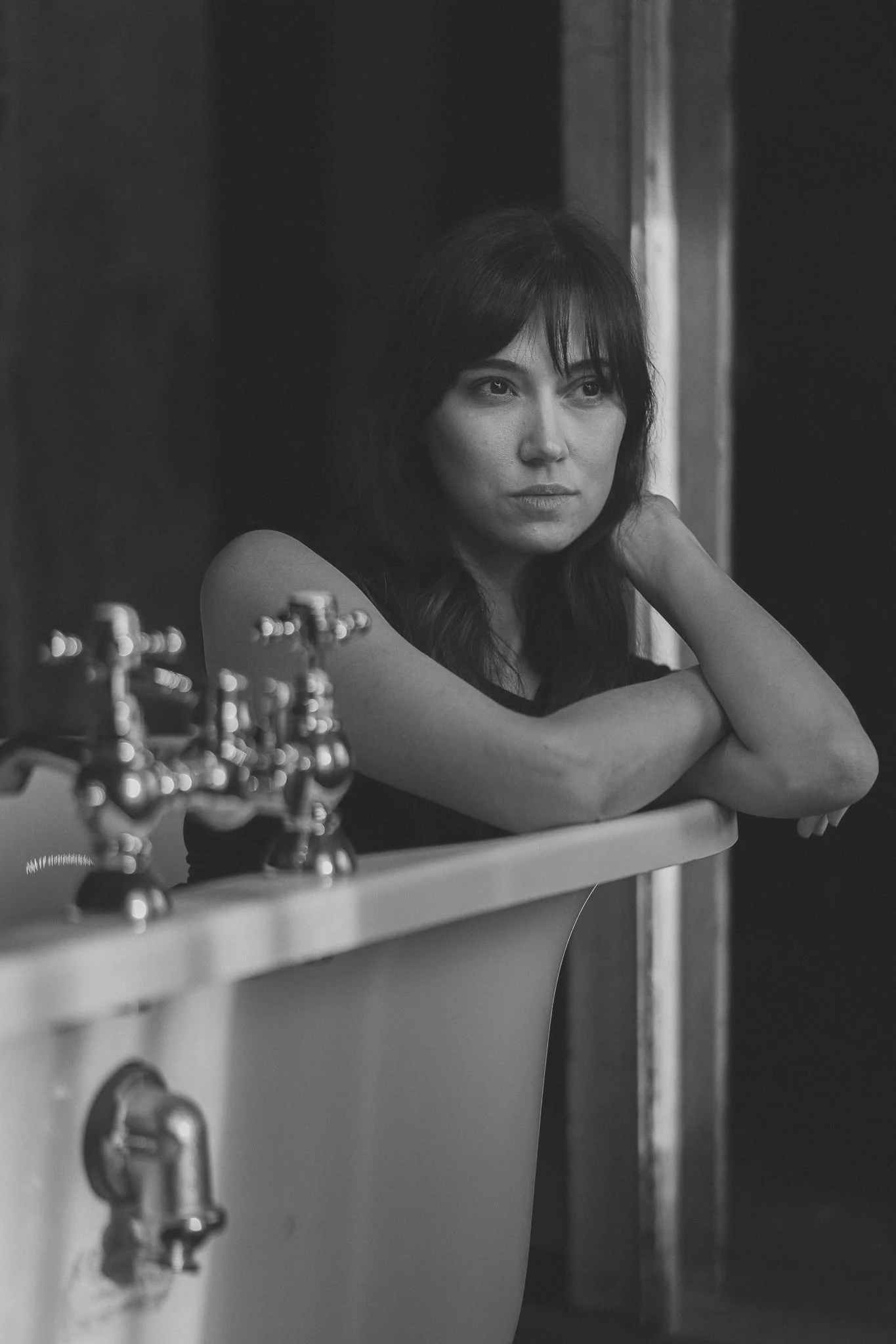 Schwarz-Weiß-Foto einer Frau mit dunklen Haaren, die an die Wand gelehnt ist, mit einem Waschbecken im Vordergrund.