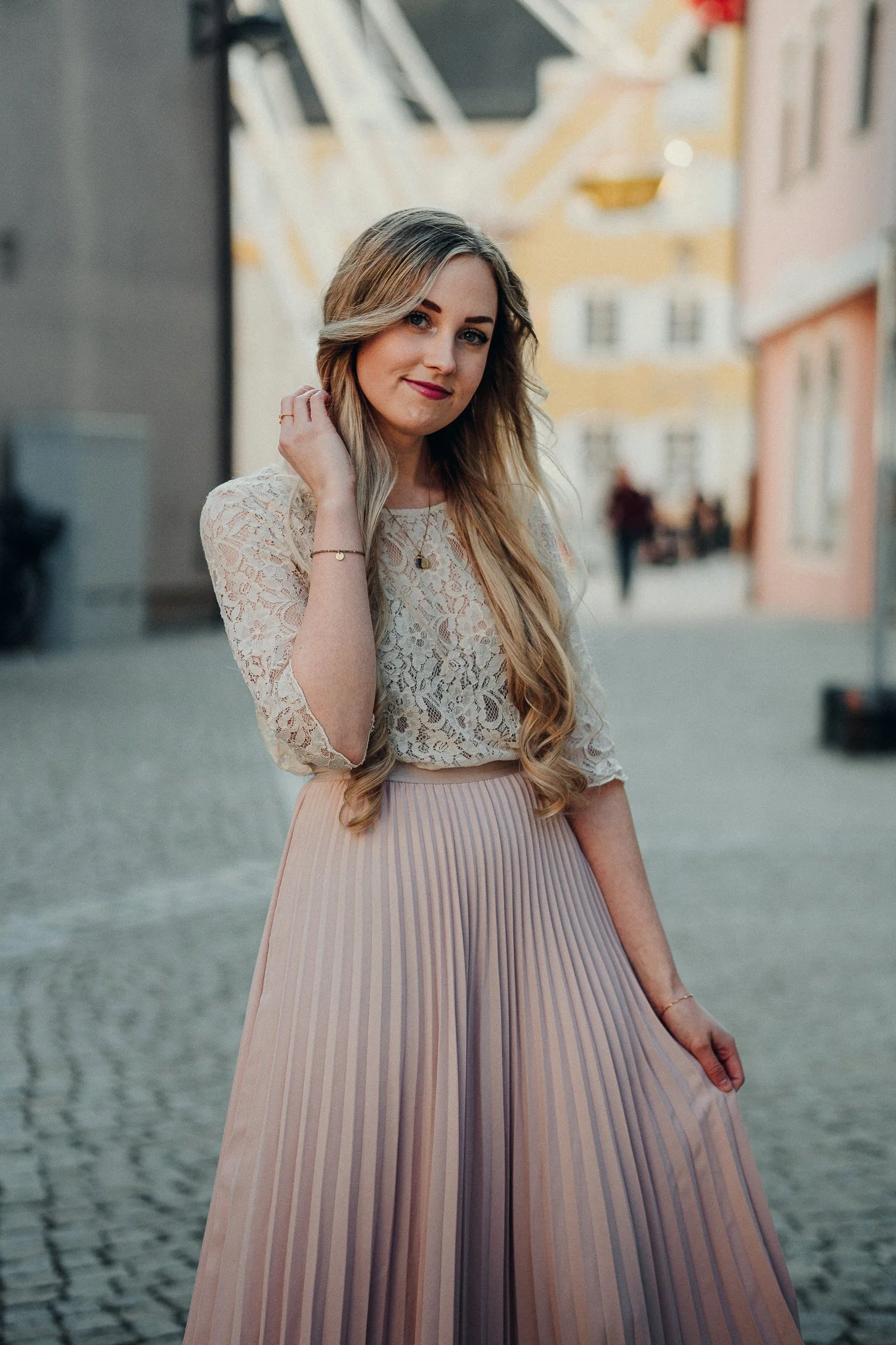 Eine junge blonde Frau in einem weißen Spitzenoberteil und einem lang gestreiften, hellrosa Rock steht auf einer Kopfsteinpflasterstraße in einer Stadt. Sie lächelt leicht und sieht in die Kamera, während sie eine Hand an den Hals legt.