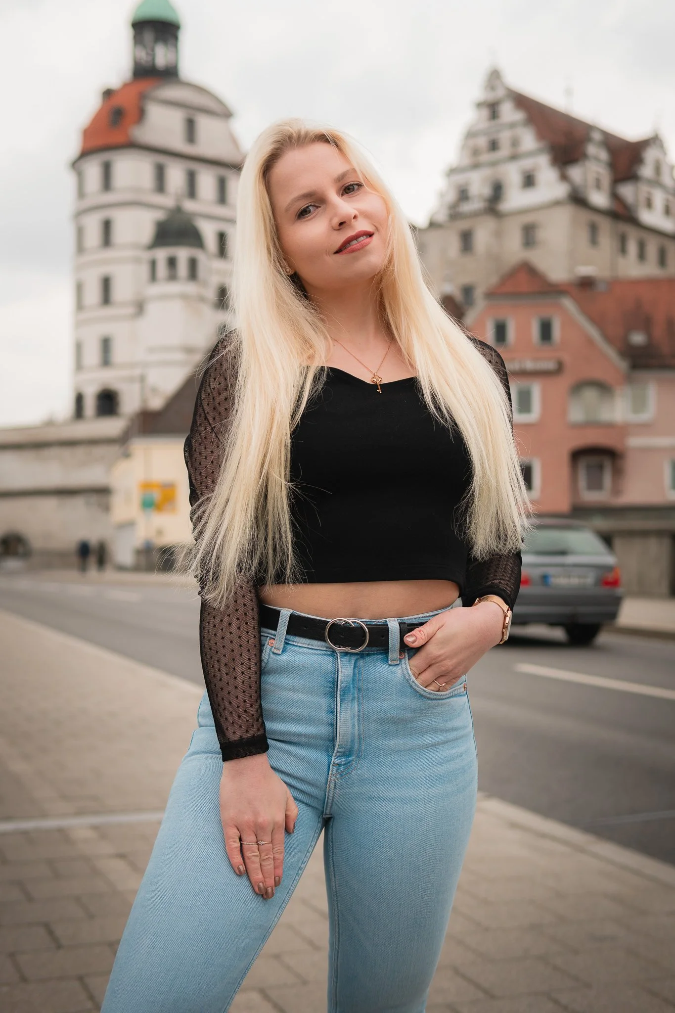 Junge blonde Frau mit schwarzem Crop-Top und hellblauen Jeans, steht auf der Straße vor historischen Gebäuden mit roten Dächern.