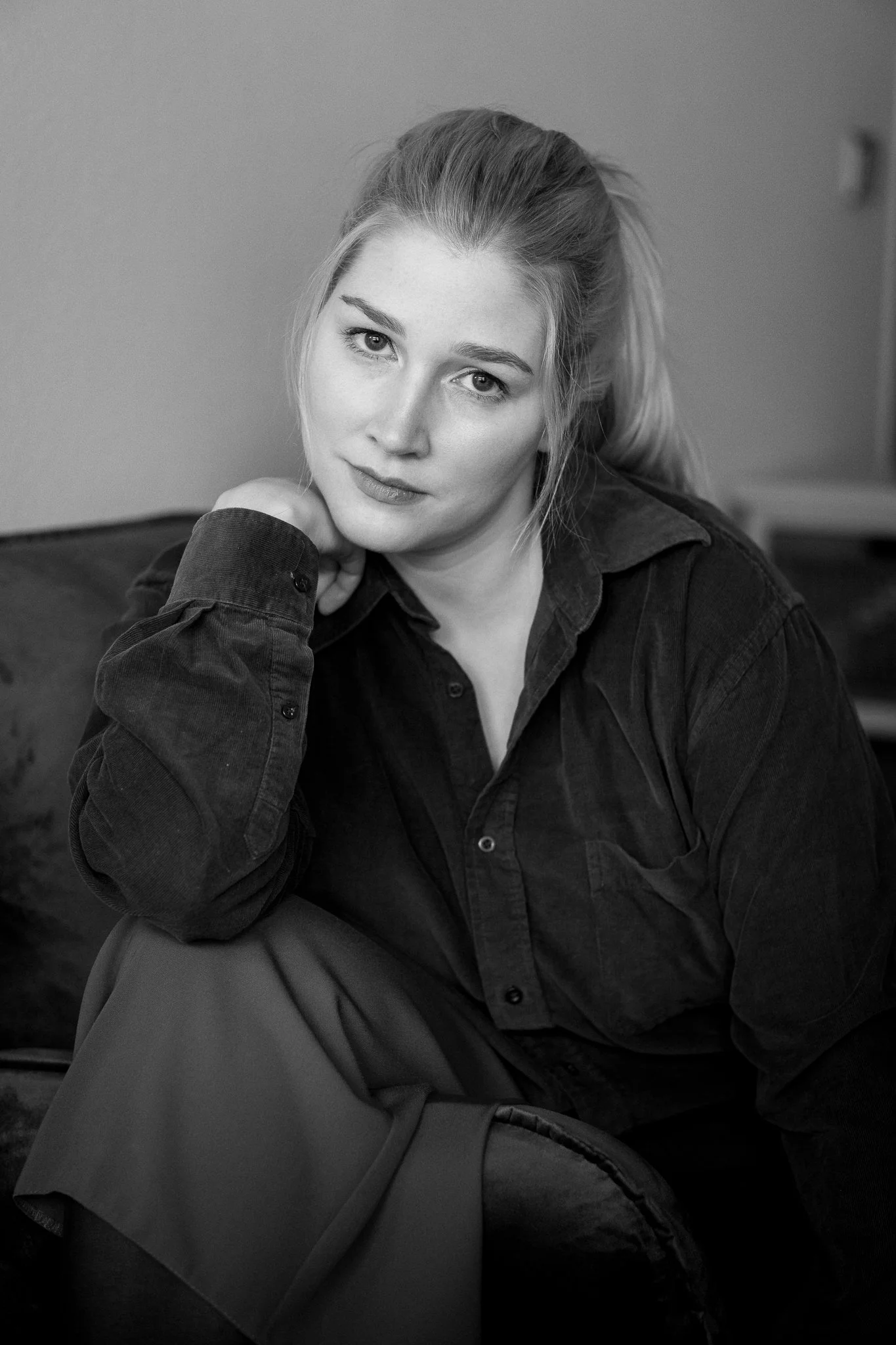 Schwarz-Weiß-Foto eines jungen Mädchens mit offener Haarfrisur, das auf einem Sofa sitzt, die Hand stützt und direkt in die Kamera schaut.