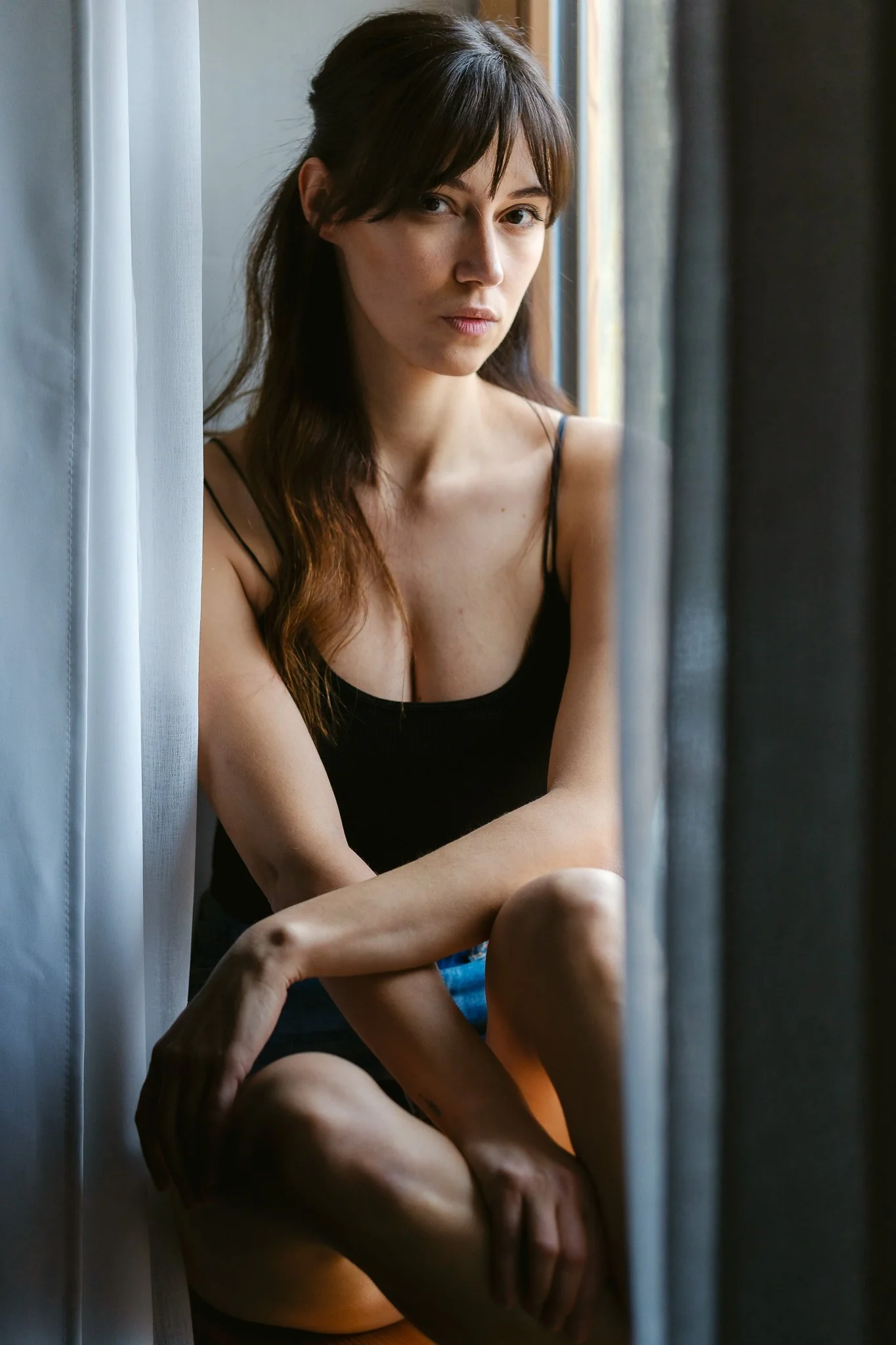 Junge Frau sitzt am Fenster, trägt ein schwarzes Top und blickt nach draußen, umgeben von Vorhängen.