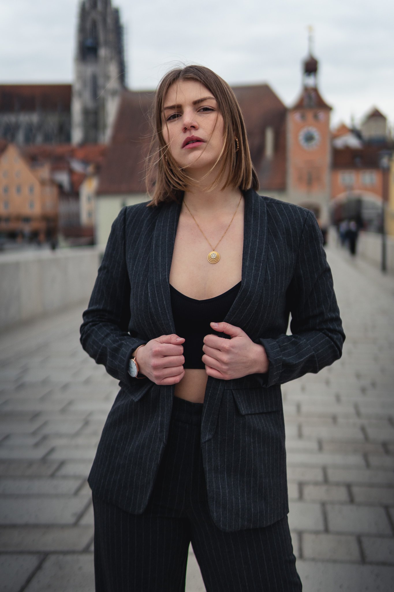 Junge Frau in schwarzem Anzug mit goldener Halskette steht im Freien auf einer Brücke, im Hintergrund sind historische Gebäude und Kirchtürme zu sehen.