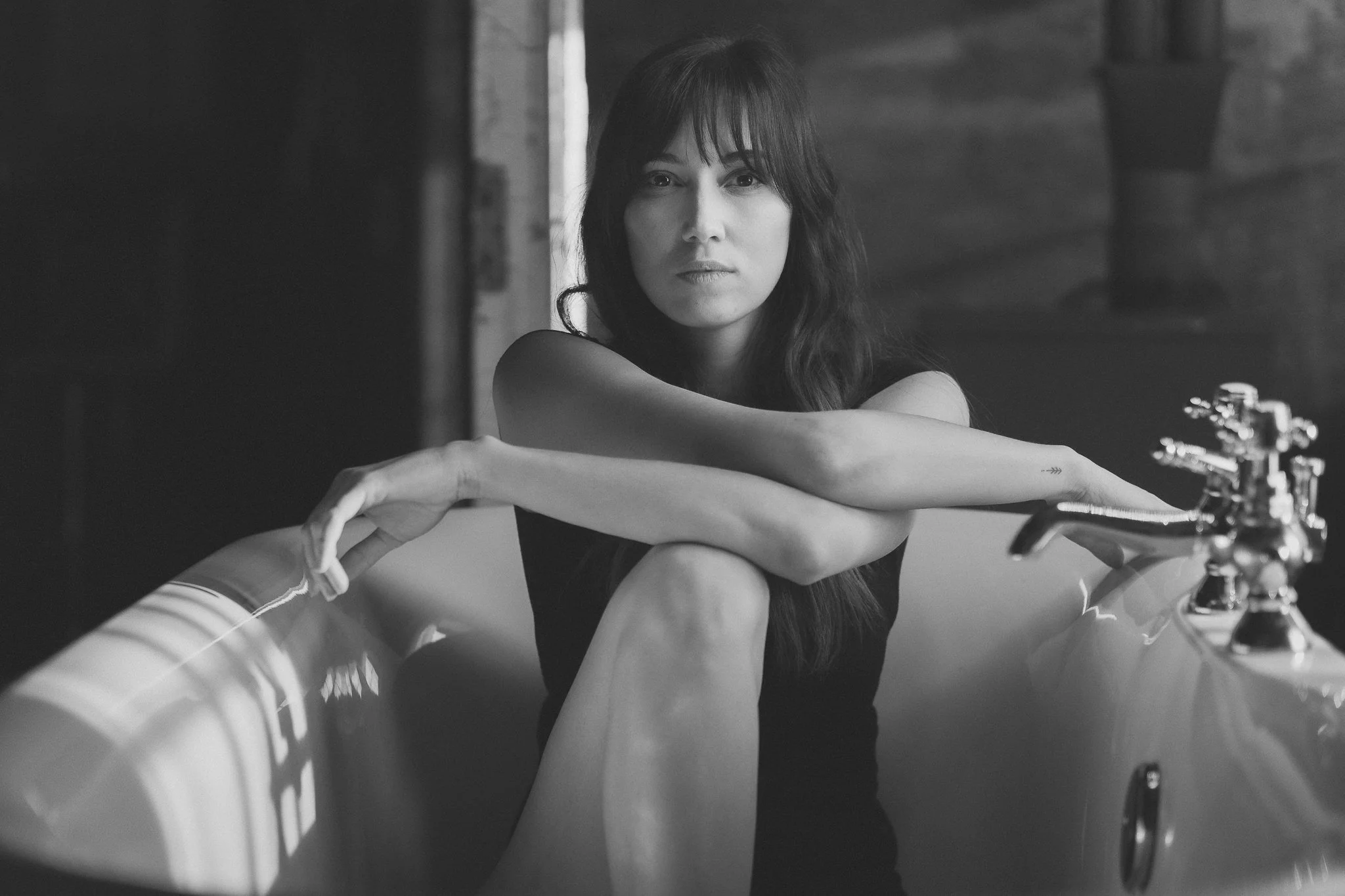 Schwarz-weiß Foto einer jungen Frau mit dunklen, langen Haaren, die in einer Badewanne sitzt, mit ihren Armen über den Knien verschränkt.