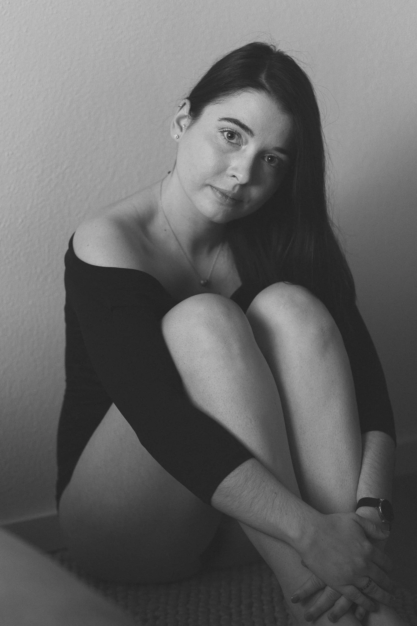 Schwarz-Weiß-Foto einer lächelnden jungen Frau mit langen dunklen Haaren, die auf dem Boden sitzt, die Knie anzieht, an die Wand gelehnt ist und Blickkontakt mit der Kamera hält.