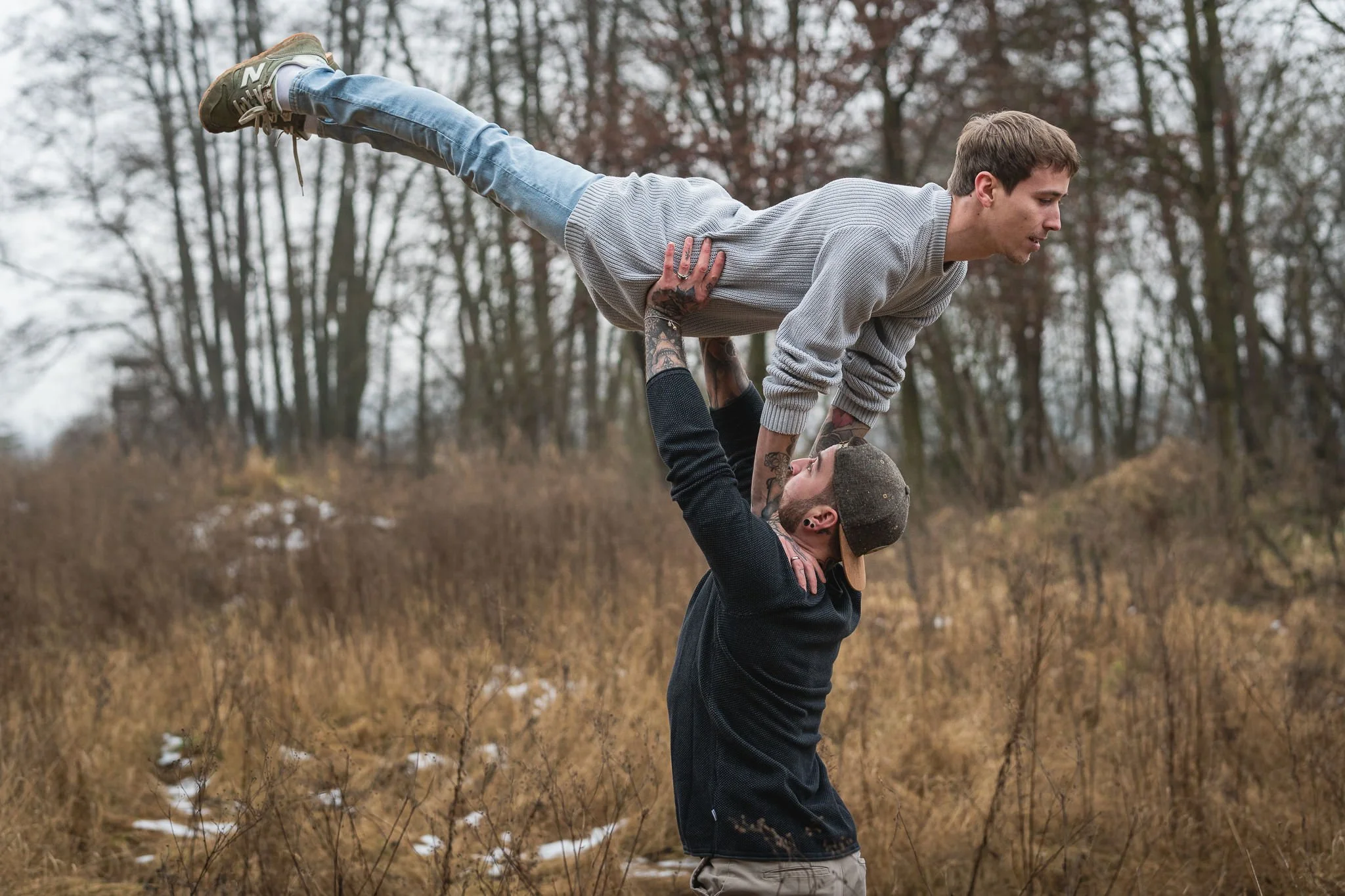 Zwei Männer, einer hält den anderen im Handstand über dem Boden, in einer Naturlandschaft mit Bäumen und trockenem Gras, bei kühlem Wetter.