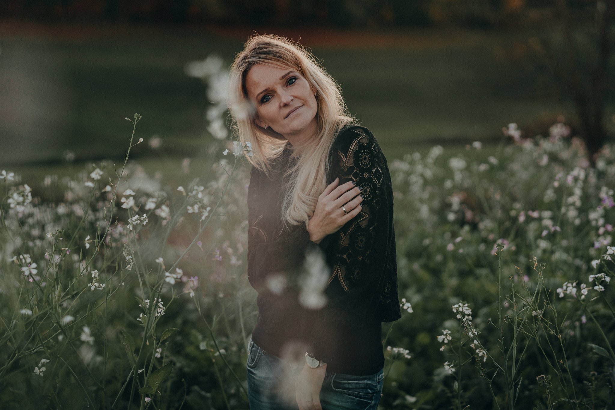 Eine blonde Frau in einem schwarzen Spitzenpullover steht in einem blühenden Feld mit weißen und lila Blumen und blickt freundlich in die Kamera.
