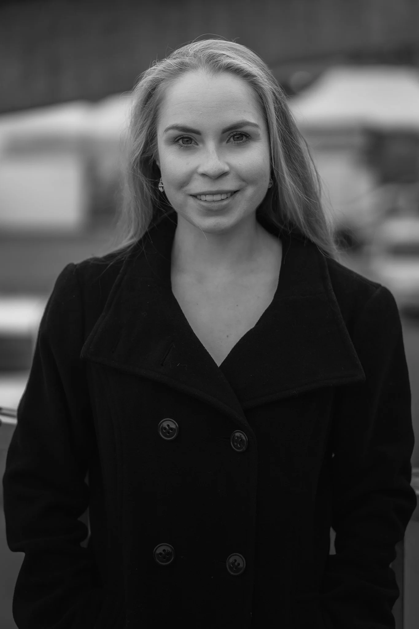 Schwarz-weiß-Foto einer lächelnden jungen Frau mit langen, glatten Haaren, die eine dunkle Jacke trägt, im Hintergrund unscharfe Außenszene.