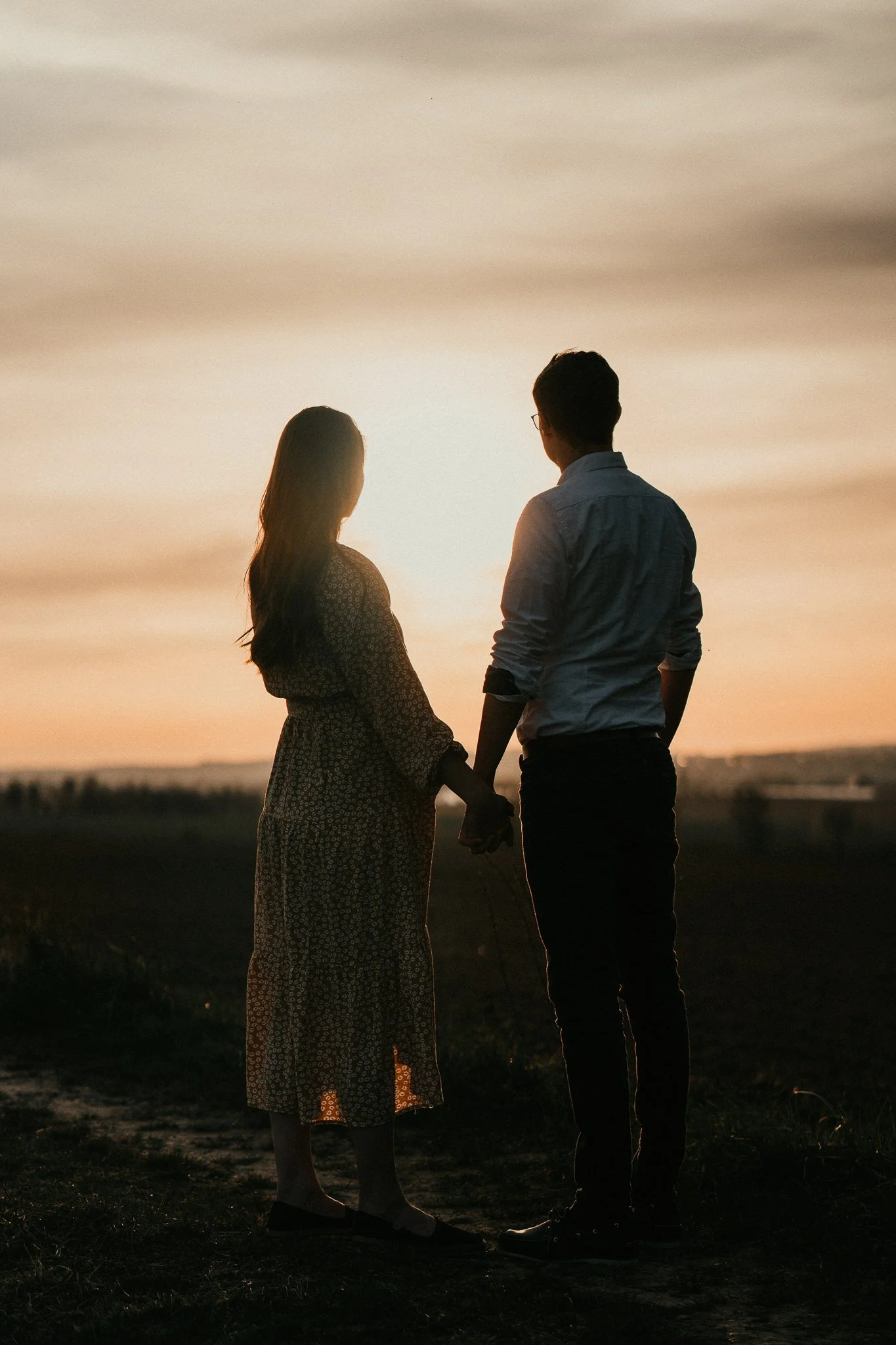 Ein Paar hält sich an einem dunklen, mit weichem Licht beleuchteten Feld bei Sonnenuntergang oder Sonnenaufgang. Sie stehen nebeneinander und schauen auf die weite Landschaft.