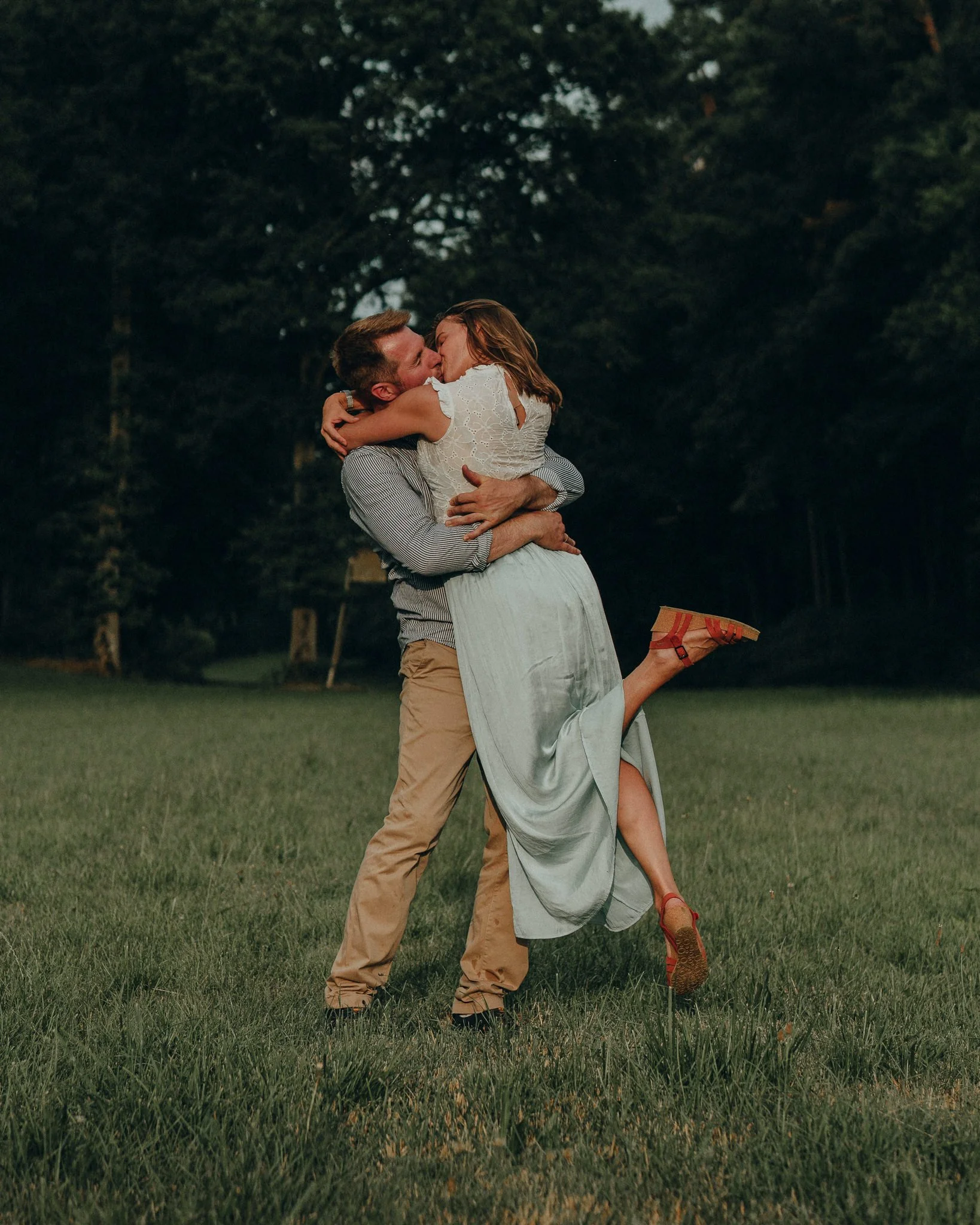 Ein Mann hebt eine Frau in einem weißen Kleid hoch, sie küssen sich liebevoll, im Hintergrund sind Bäume und Gras. Es ist Abend oder Dämmerung.