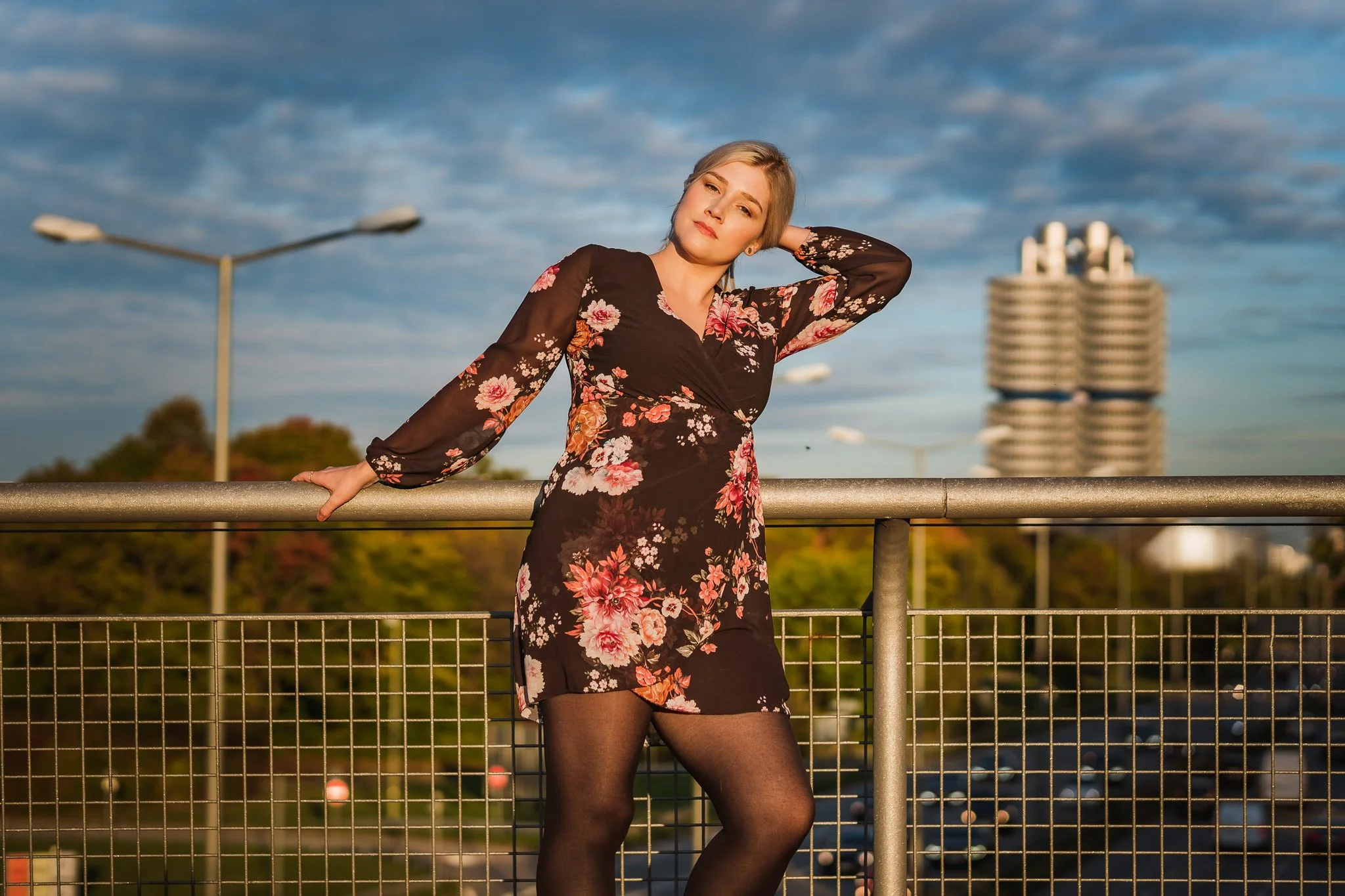 Eine junge Frau trägt ein schwarzes Kleid mit rosa und weißen Blumendrucken und schwarze Strumpfhosen. Sie steht an einer Metallbrüstung auf einer Brücke bei Sonnenuntergang, die Landschaft und ein Gebäude im Hintergrund zu sehen. Sie hat blondes, sc