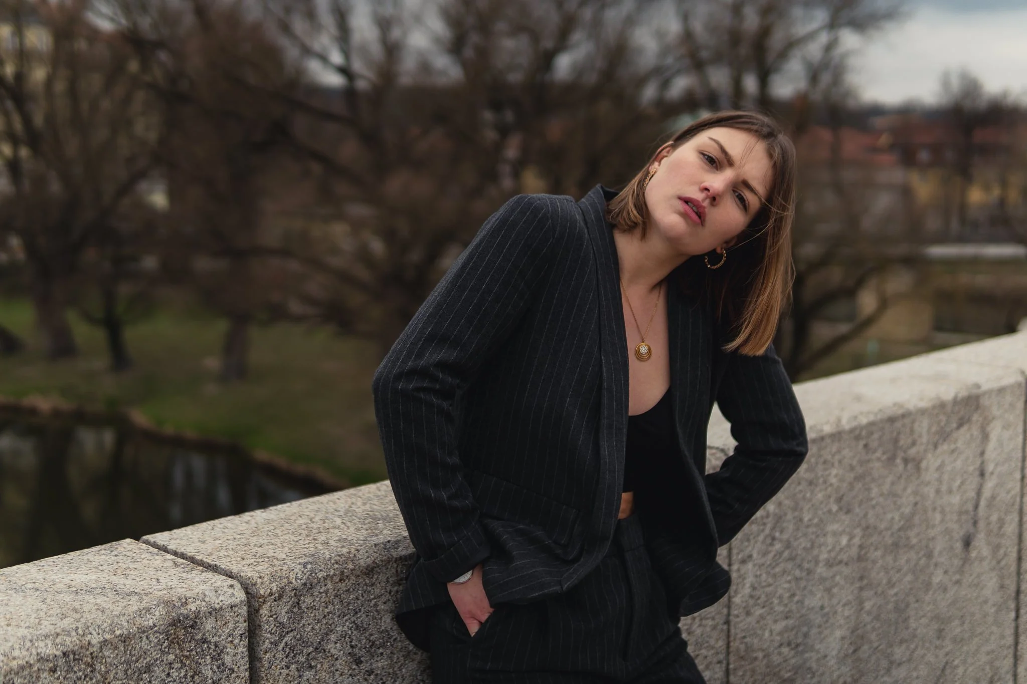 Junge Frau in dunklem Anzug lehnt an einer Steinmauer im Freien bei bewölktem Himmel, mit Bäumen im Hintergrund.