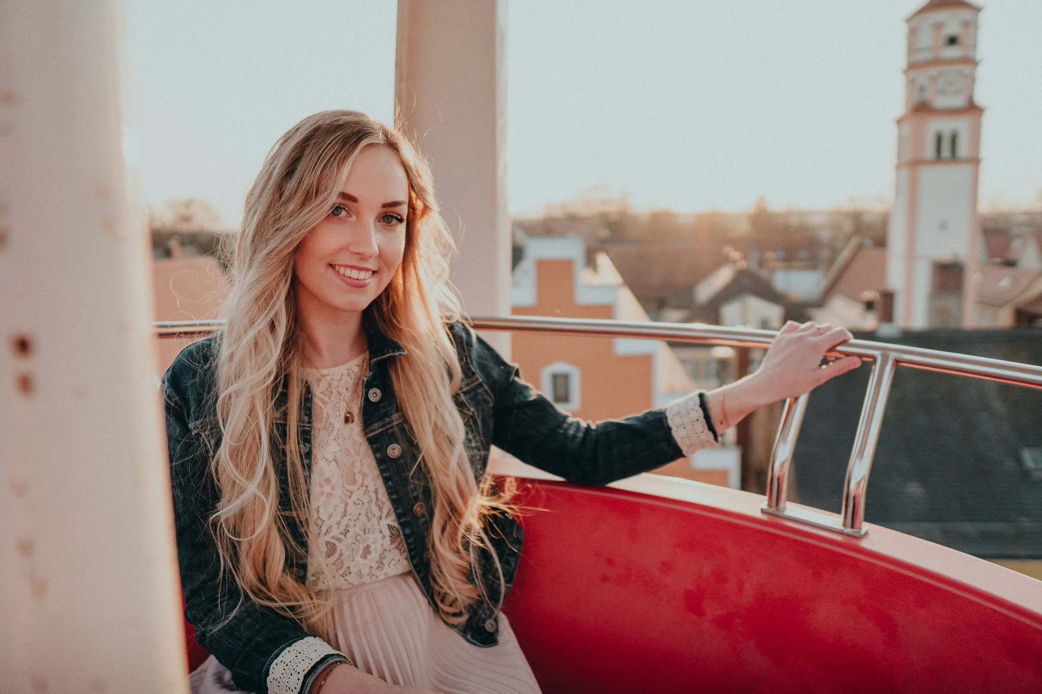 Eine junge Frau mit langem, lockigem blondem Haar sitzt in einer Riesenradfahrkabine bei Sonnenuntergang, trägt eine dunkle Jeansjacke, ein weißes Spitzenkleid, und lächelt zur Kamera.