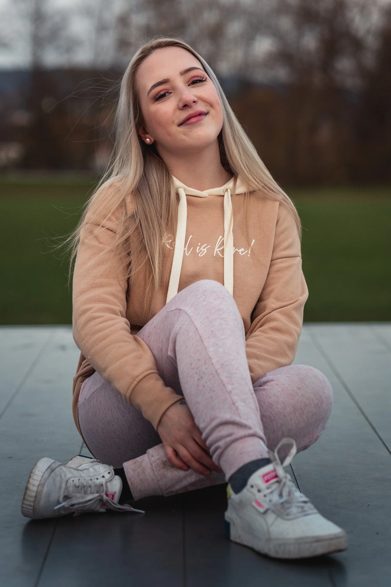 Junge Frau sitzt im Freien auf einem dunklen Boden, trägt einen beigen Hoodie, pinkfarbene Leggings, weiße Turnschuhe, lächelt und blickt zufrieden in die Kamera.