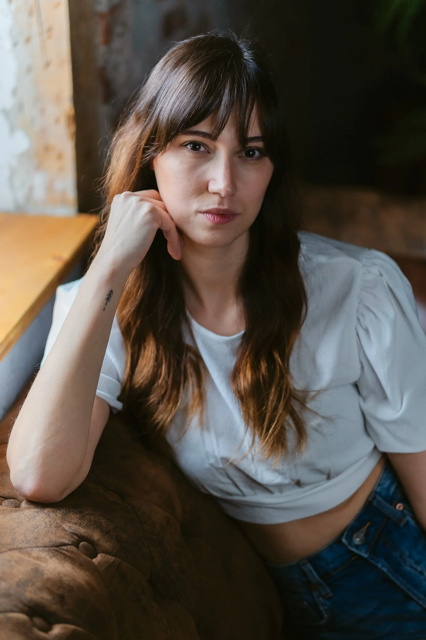 Eine junge Frau mit braunen Haaren und einem weißen T-Shirt sitzt auf einem braunen Ledersofa in einem rustikalen Raum und blickt direkt in die Kamera.