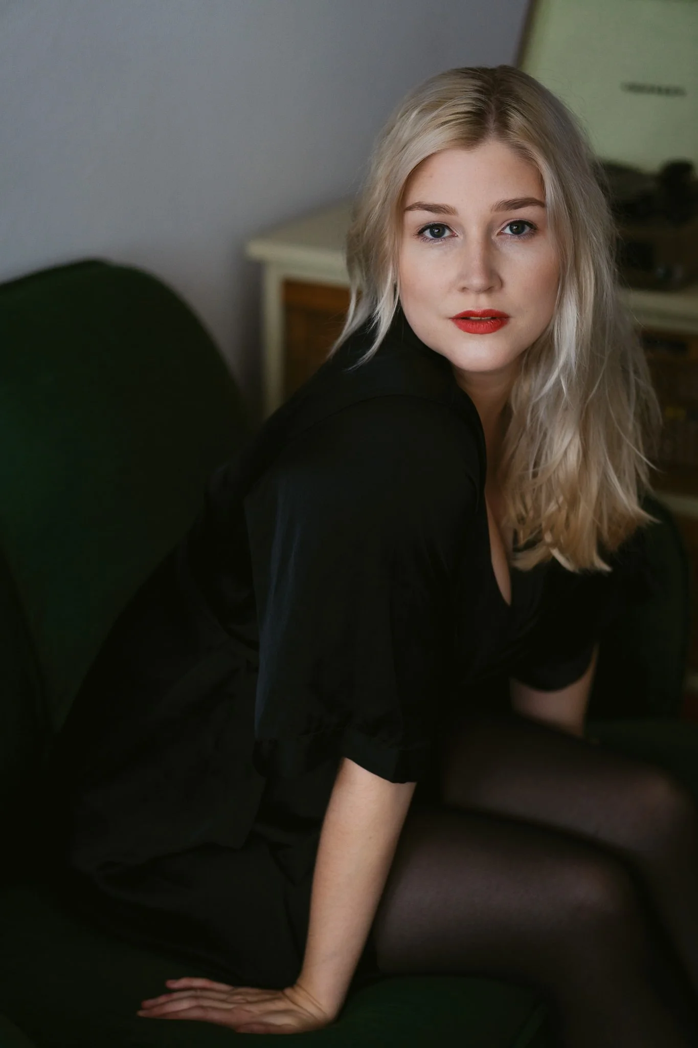 Junge Frau mit blondem, lockigem Haar, rotem Lippenstift und schwarzer Kleidung sitzt auf einem grünen Sofa in einem Raum mit grauer Wand und einem kleinen Tisch im Hintergrund.