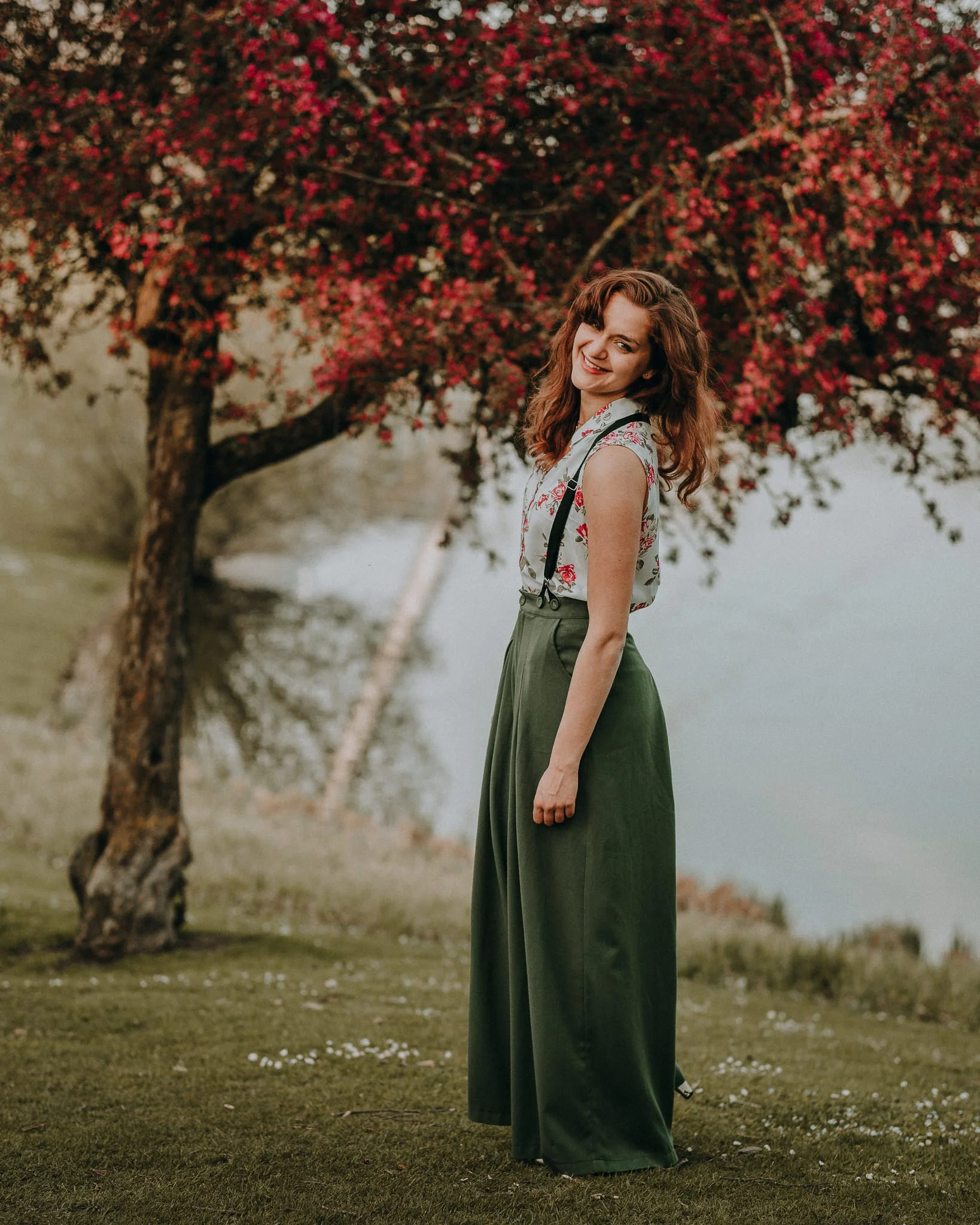Eine junge Frau steht im Grünen vor einem Baum mit rosa Blüten, lächelt und schaut in die Kamera.