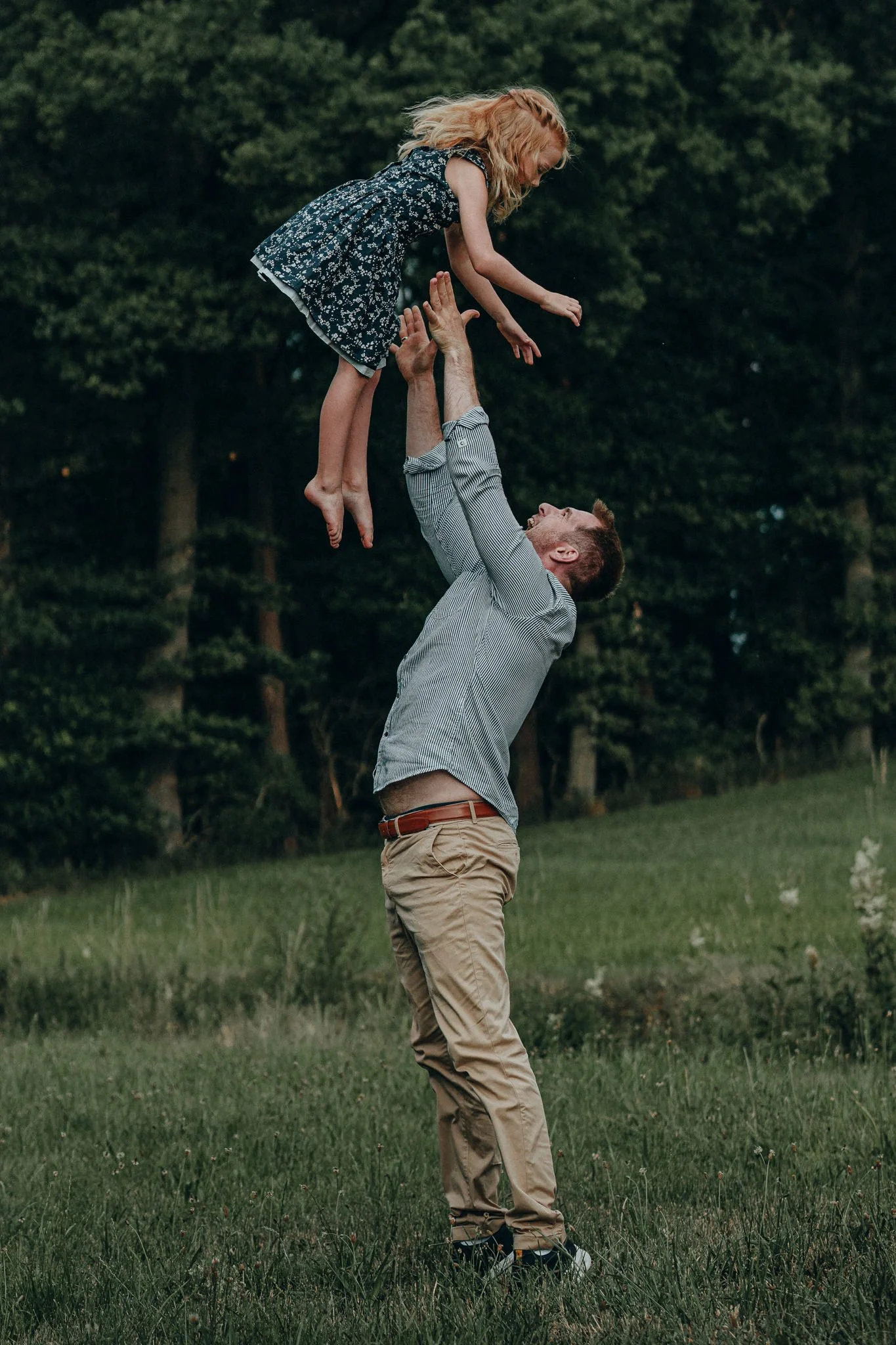 Vater wirft Tochter lachend in die Luft im Freien, mit Bäumen im Hintergrund.