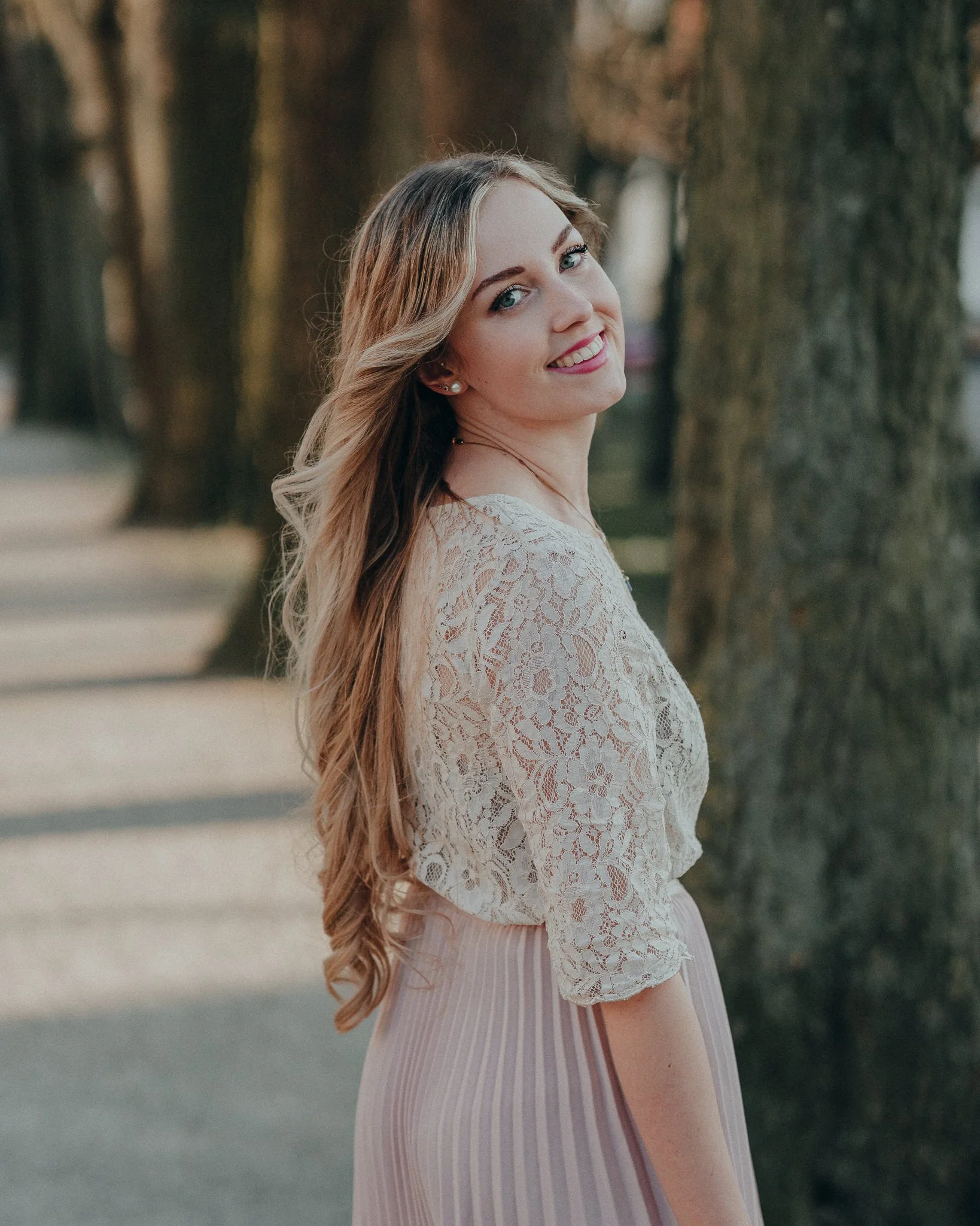 Eine lächelnde junge Frau mit langen, gewellten Haaren steht vor Bäumen im Freien, trägt ein weißes Spitzenoberteil und einen hellrosa, gestreiften Rock.