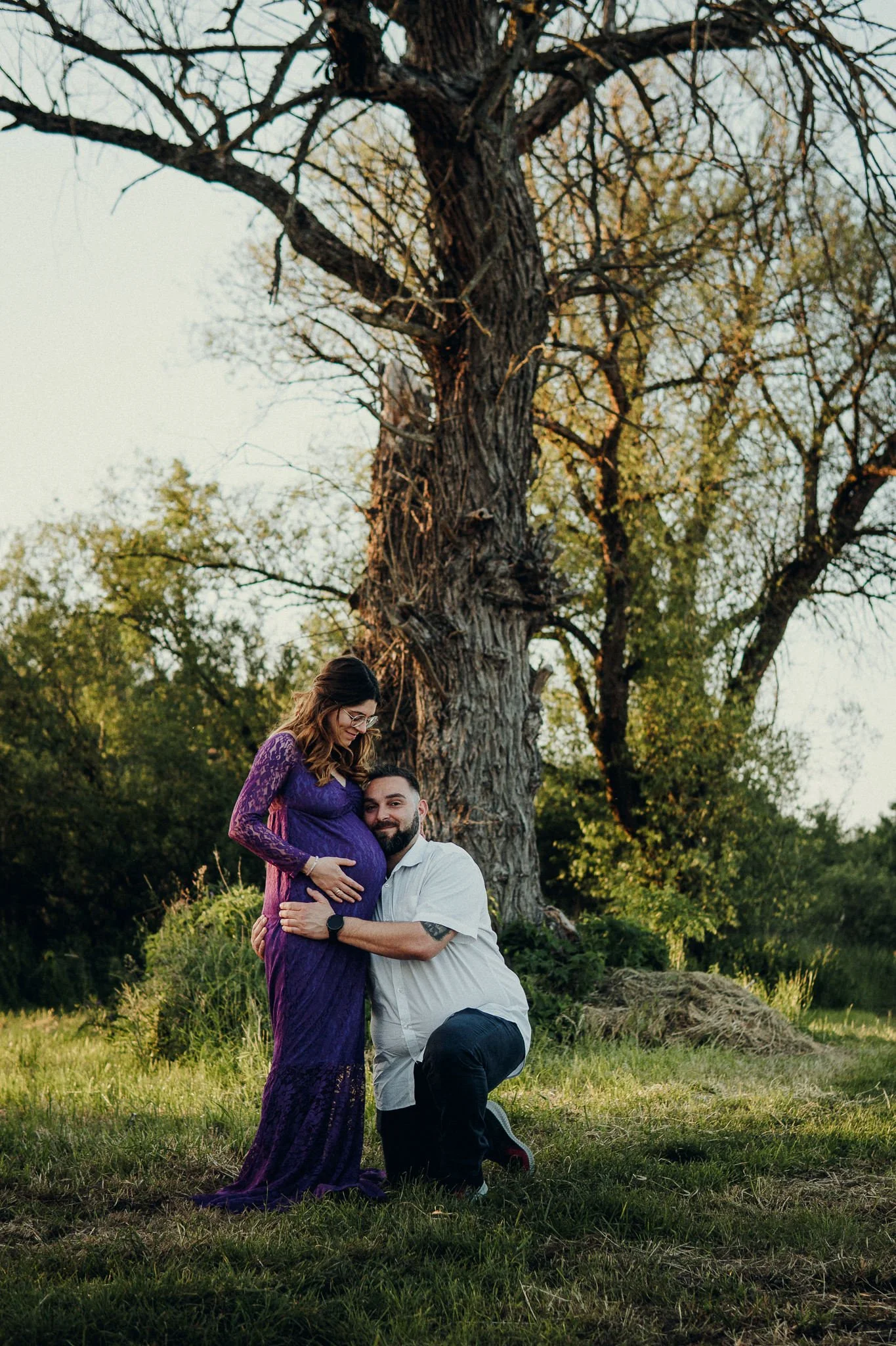 Ein schwangertes Paar vor einem Baum im Freien, Frau trägt ein lila Kleid, er trägt ein weißes Hemd und dunkle Hosen, sie umarmen sich liebevoll.