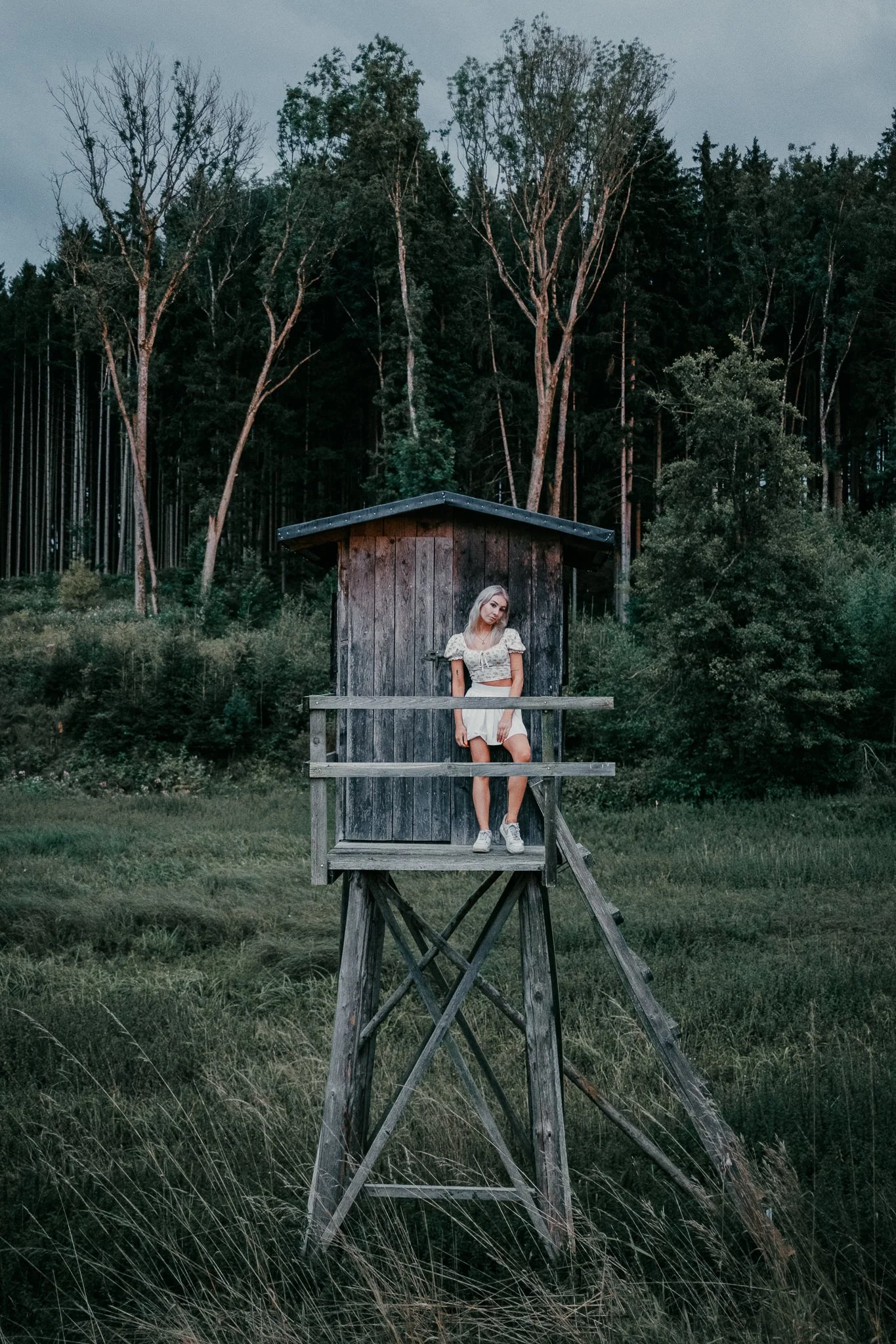 Junge Frau steht auf einer Baumhaus-Terrasse in einer waldähnlichen Umgebung, umgeben von Bäumen und Grünflächen.