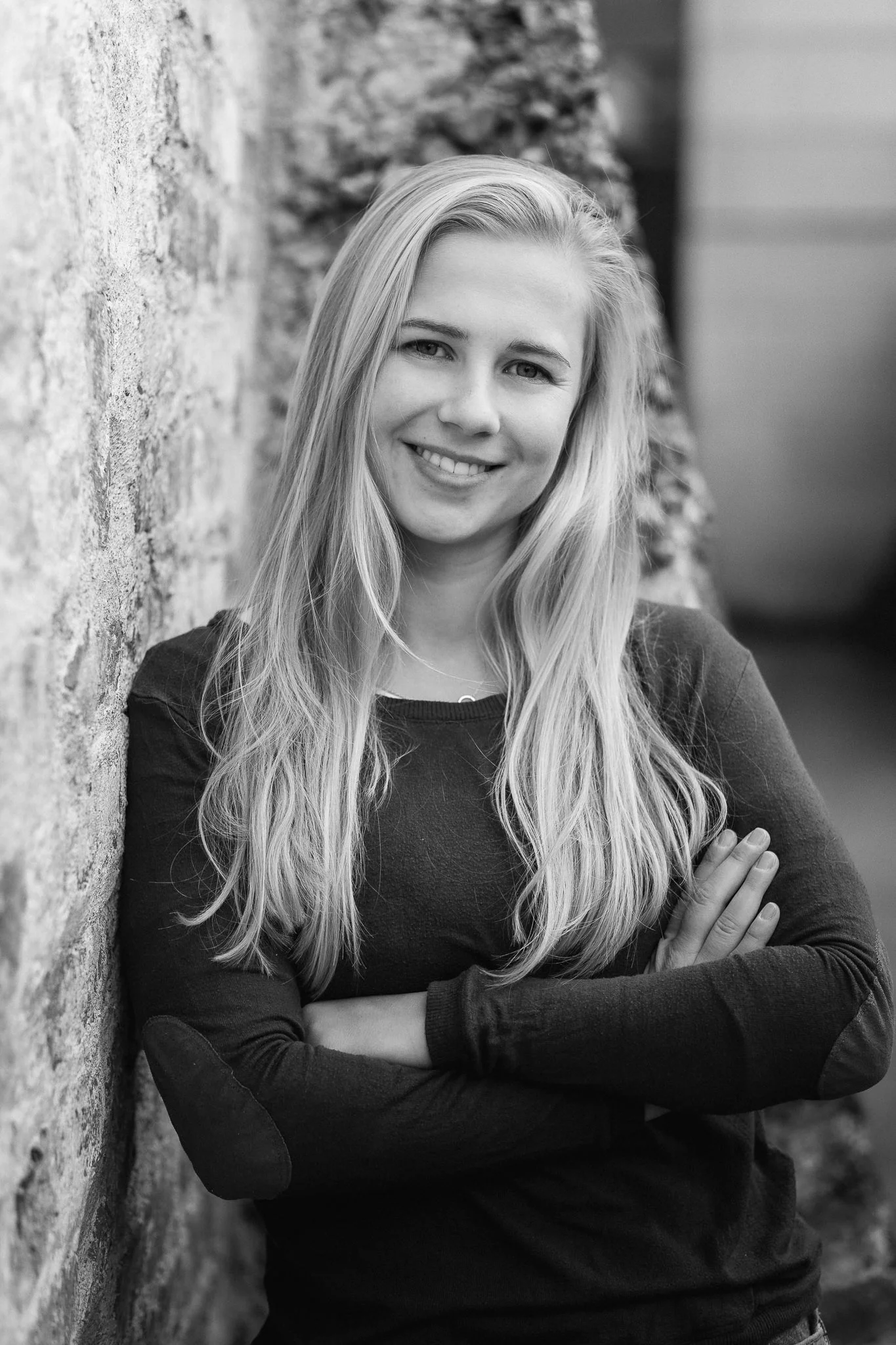 Schwarz-weiß Porträtaufnahme einer jungen Frau mit langen, lockigen Haaren, die vor einer Steinmauer lächelnd mit verschränkten Armen steht.
