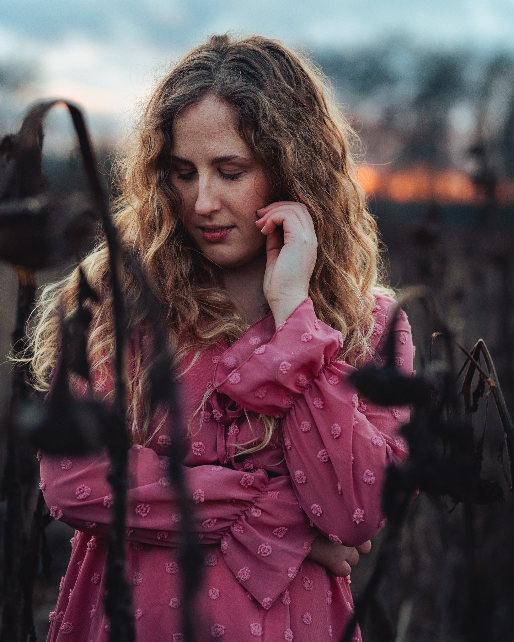 Junge Frau mit lockigem Haar trägt ein pinkfarbenes Kleid mit kleinen Blumenmustern, steht in einem Feld bei Sonnenuntergang, blickt nach unten, mit nachdenklicher Miene.