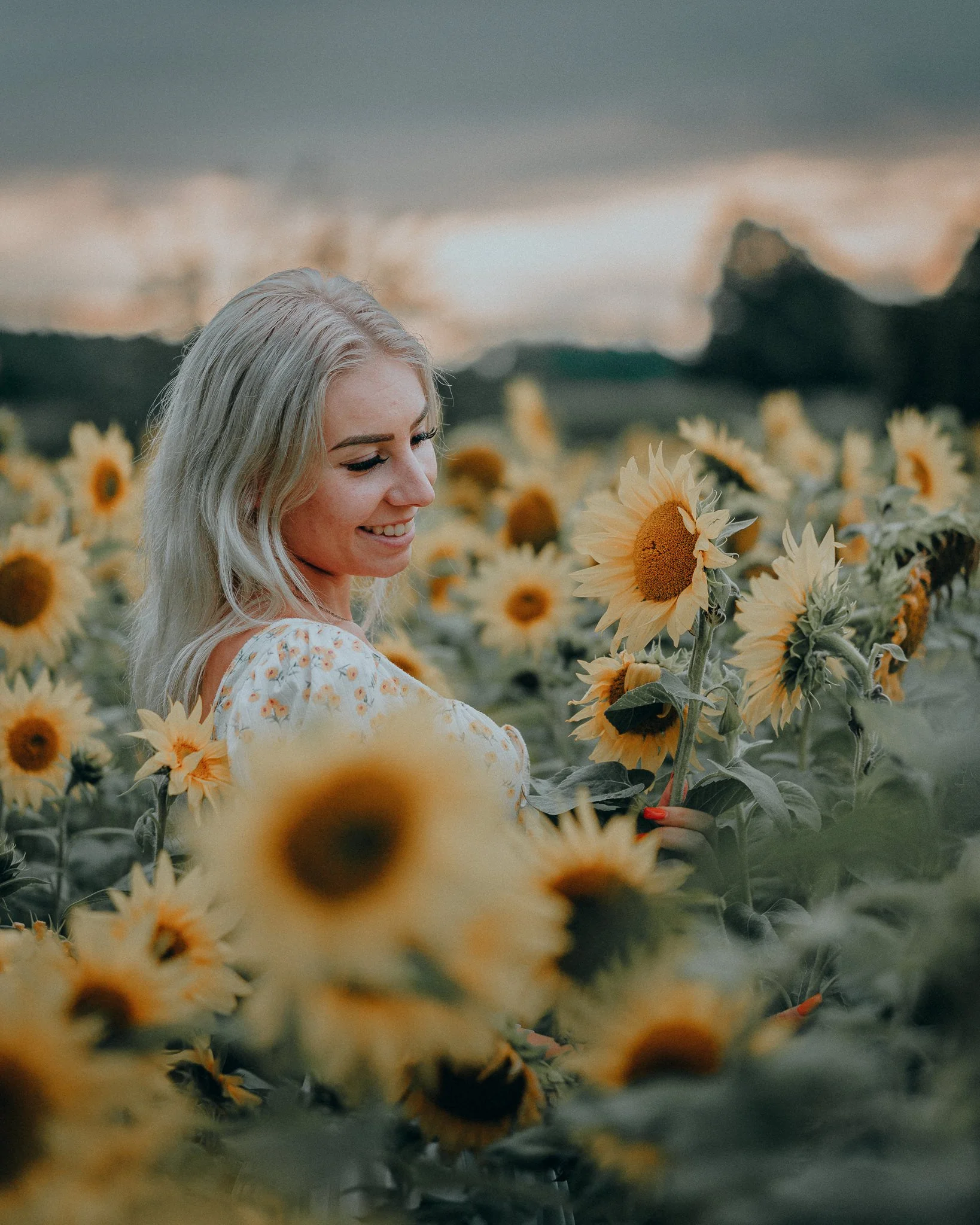Eine lachende junge Frau mit blonden Haaren steht in einem Sonnenblumenfeld bei Sonnenuntergang, umgeben von gelben Sonnenblumen.