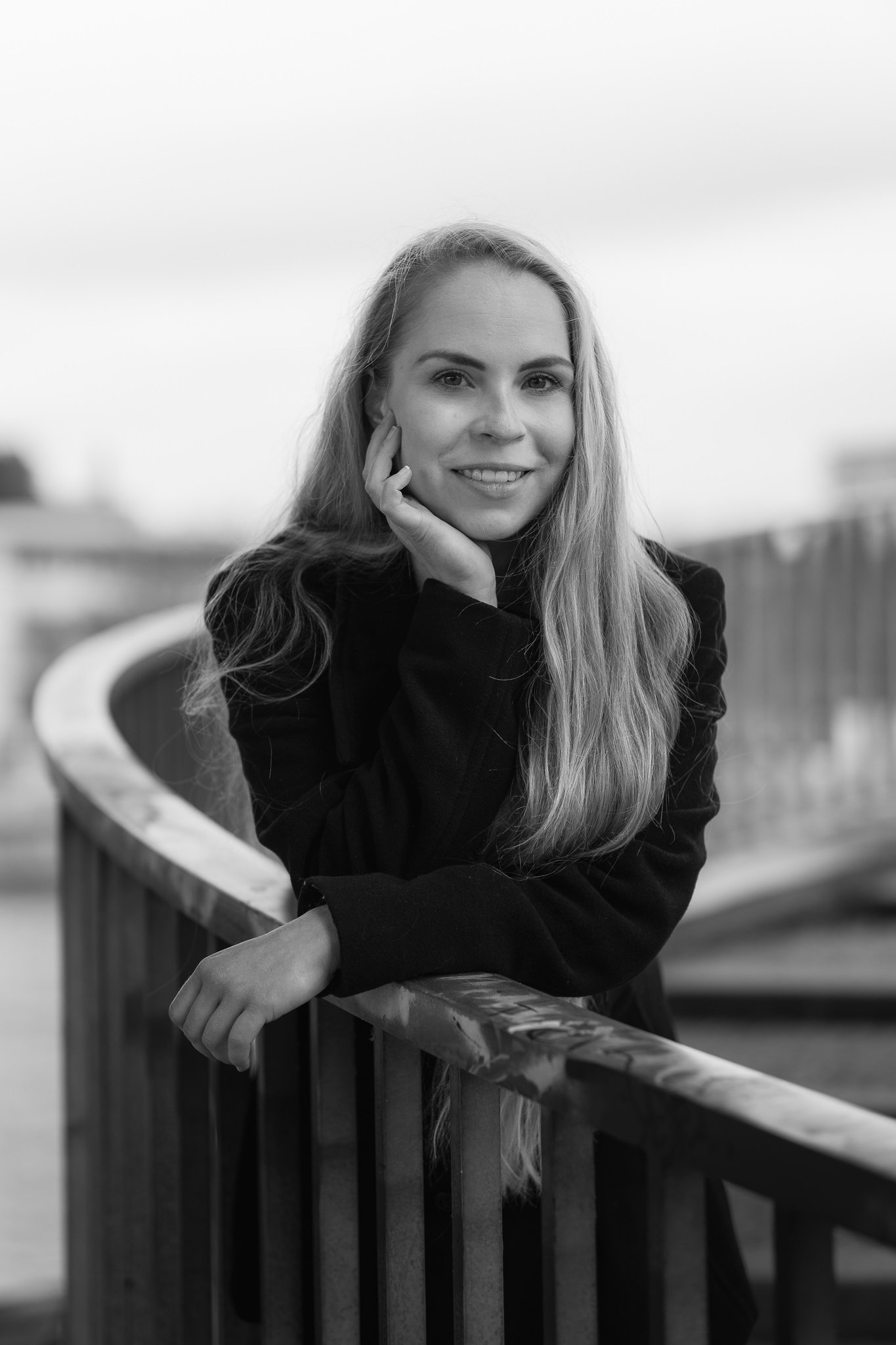 Schwarz-weiß-Fotografie einer lächelnden jungen Frau, die auf einem Brückenrand lehnt, mit langem Haar, draußen vor einem urbanen Hintergrund.