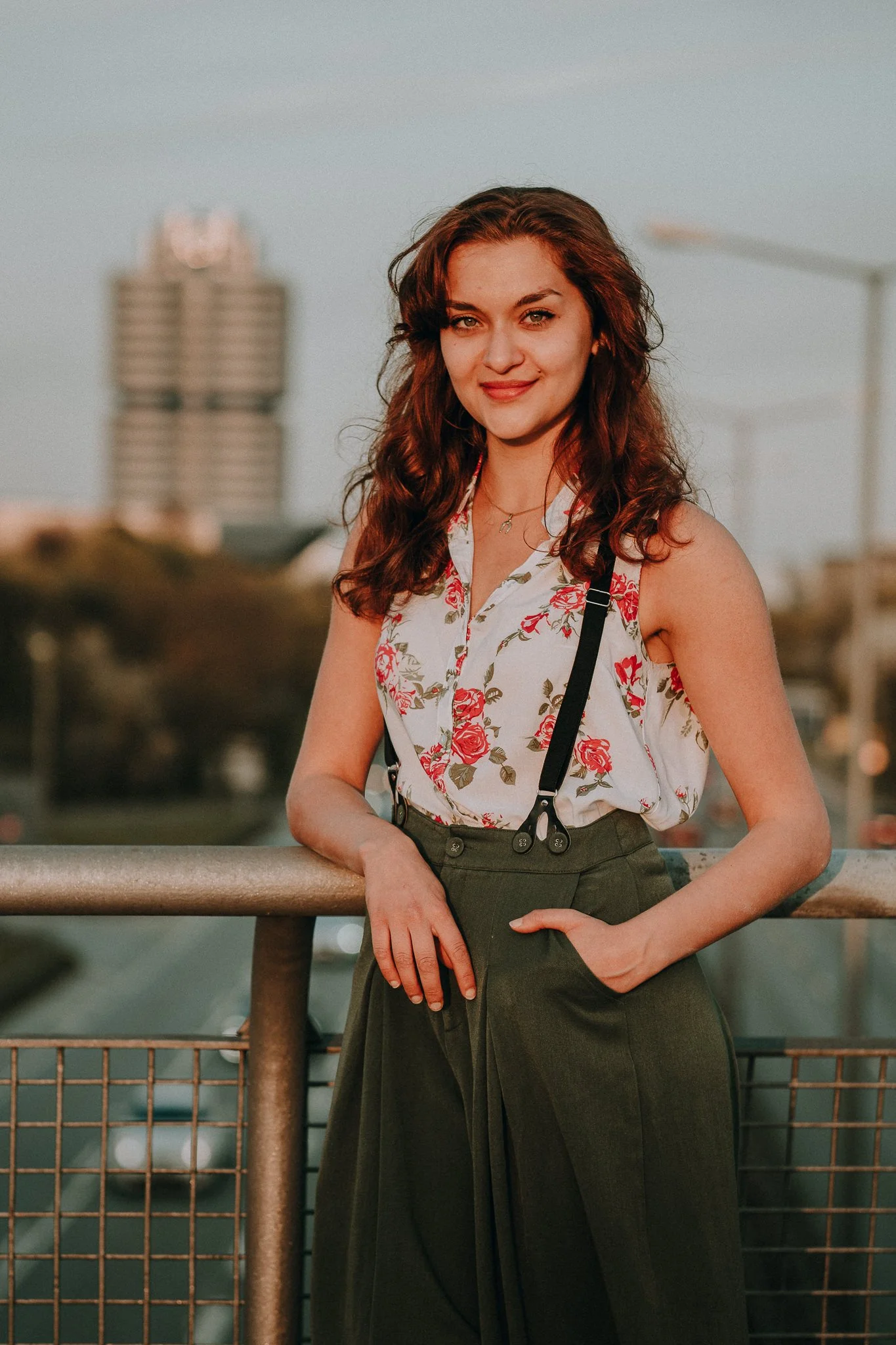 Junge Frau mit braunen lockigen Haaren trägt ein weißes Hemd mit roten Blumen, steht an einem Geländer bei Sonnenuntergang, im Hintergrund sind Gebäude und ein Himmel sichtbar.