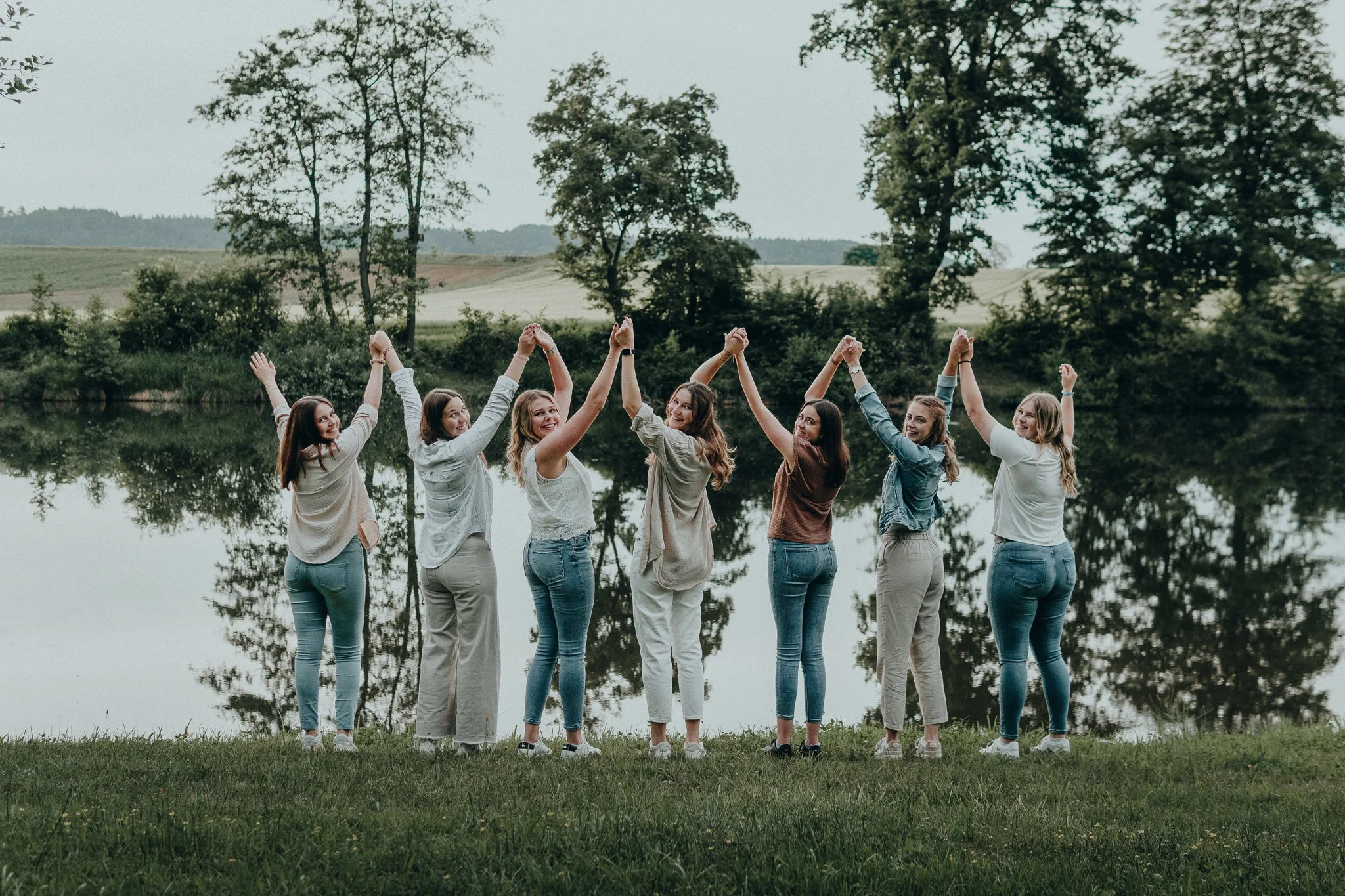 Sieben junge Frauen stehen am Ufer eines Sees, halten sich an den Händen und heben die Arme in die Luft, während sie sich lächelnd anschauen, mit Bäumen im Hintergrund.