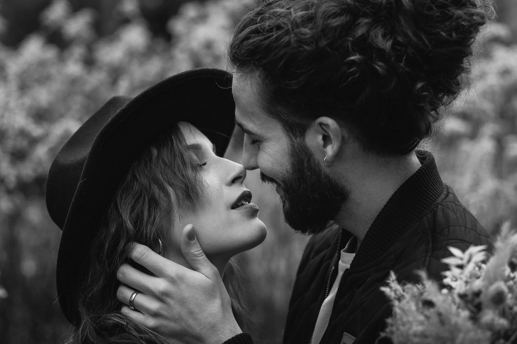 Ein Mann und eine Frau stehen sich nah beieinander, fast mit ihren Nasen berührend, im Freien mit unscharfem Hintergrund. Der Mann hat dunkles, lockiges Haar, einen Bart und ein Ohrring, die Frau trägt einen Hut und hat lange, lachende Augen.
