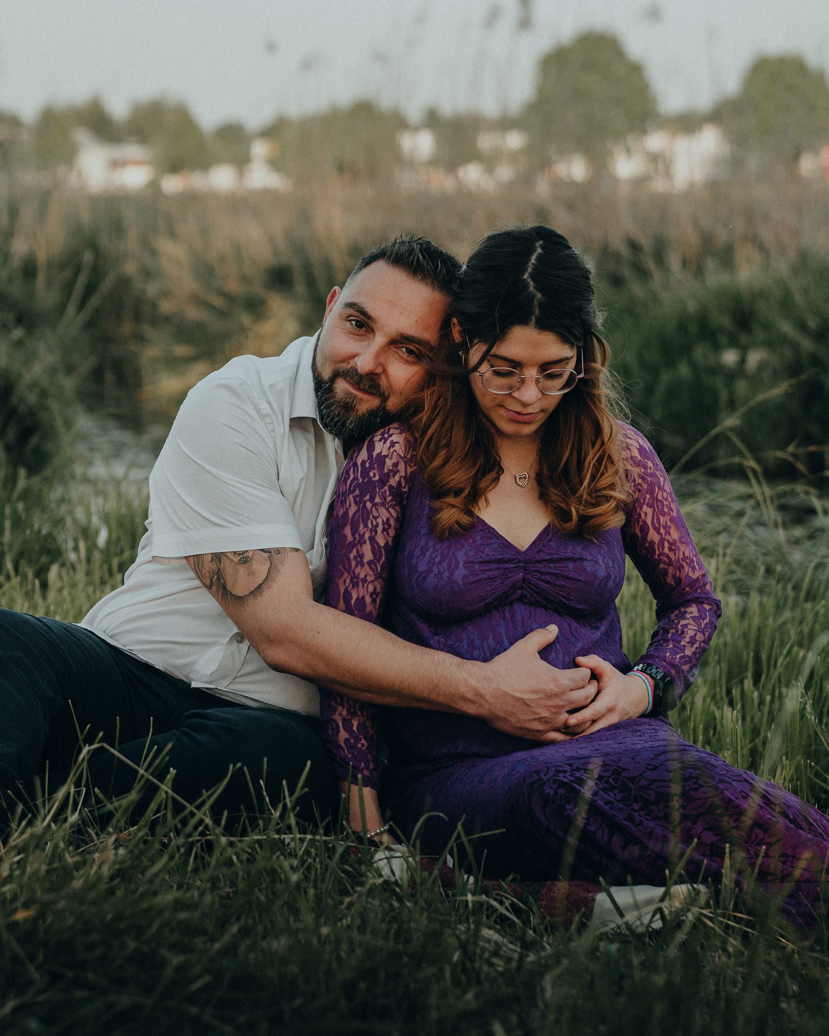 Ein Paar sitzend im Freien auf Gras, umarmt, die Frau ist schwanger, die Männer umarmt die Frau auf den Bauch