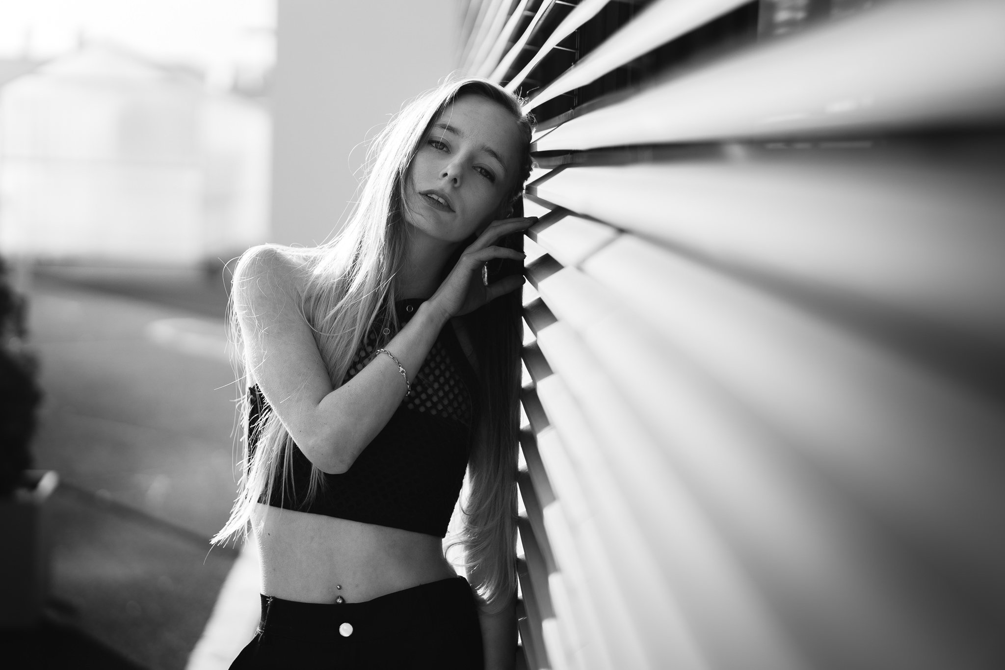 Schwarz-Weiß-Foto einer jungen Frau mit langen Haaren, die an einer horizontalen Wand lehnt und direkt in die Kamera schaut.