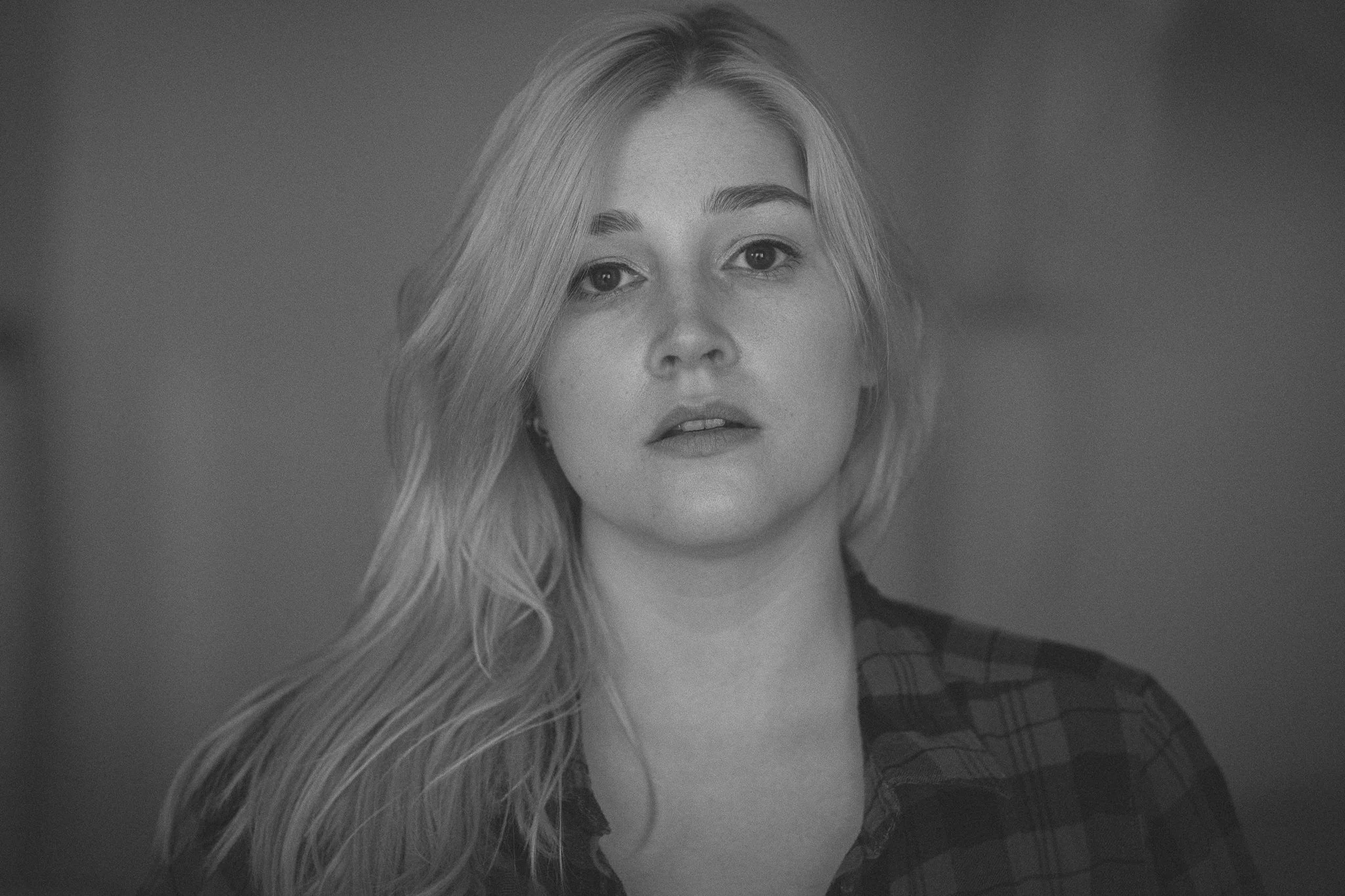 Schwarz-Weiß-Porträt einer jungen Frau mit langen, gewellten Haaren, die in eine Richtung fallen, und einem karierten Hemd, die leicht den Kopf zur Seite neigt.