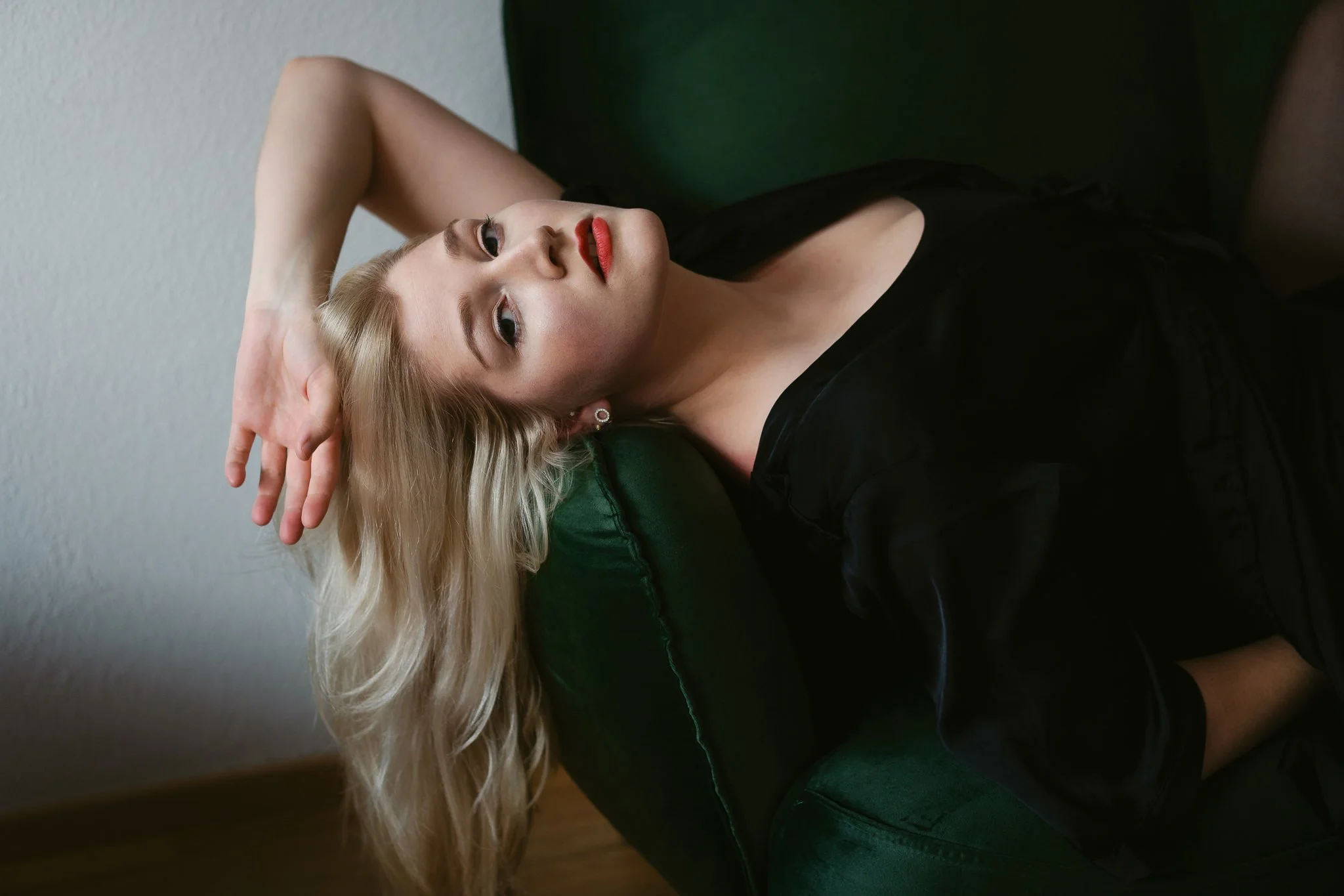 Junge Frau mit lange blondem Haar liegt auf einer grünen Couch, trägt schwarzes Kleid, rote Lippen, blickt nach oben, Hand im Haar.
