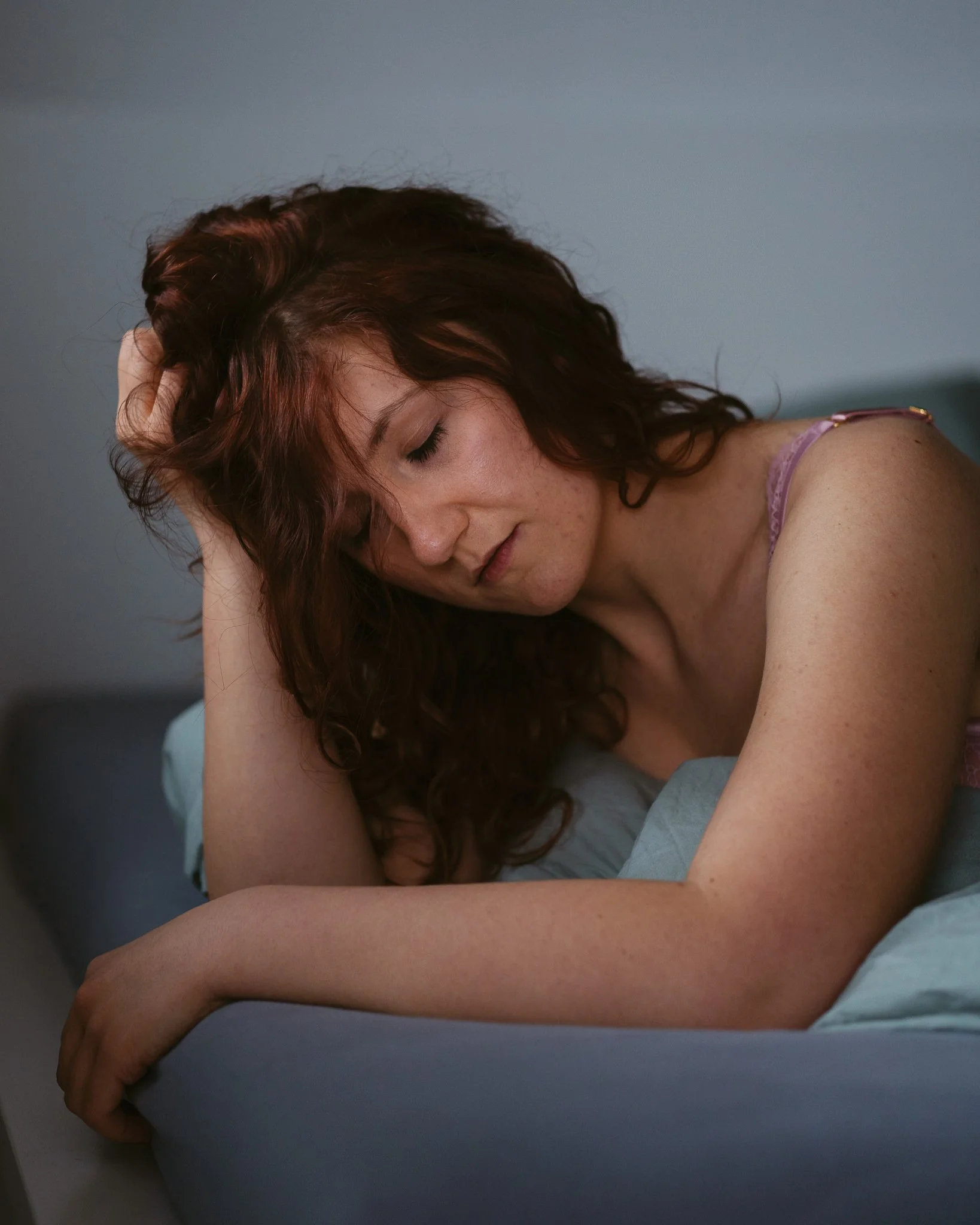 Junge Frau mit lockigem roten Haar, die auf einem Bett mit geschlossenen Augen sitzt, den Kopf auf die Hand gestützt und nachdenklich wirkt.