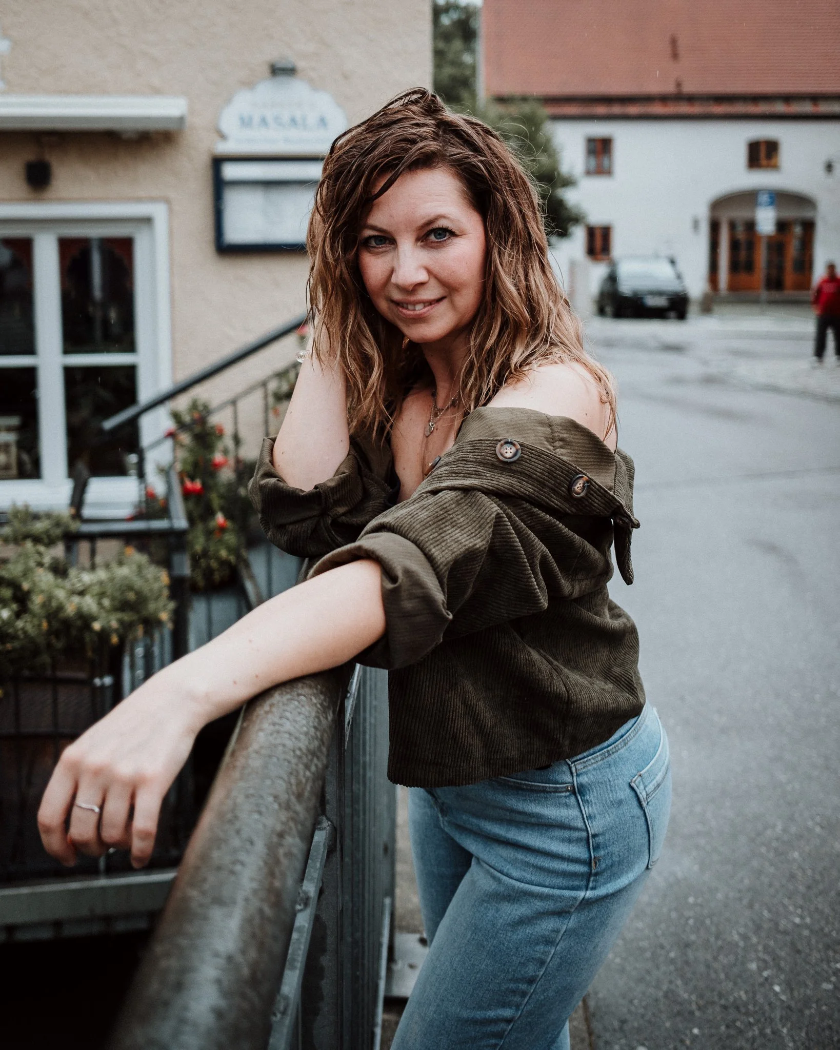 Junge Frau mit braunen, lockigen Haaren, die auf einer Brüstung in einer Straße in Deutschland lehnt, trägt eine off-shoulder Jacke und eine Halskette, lächelt.