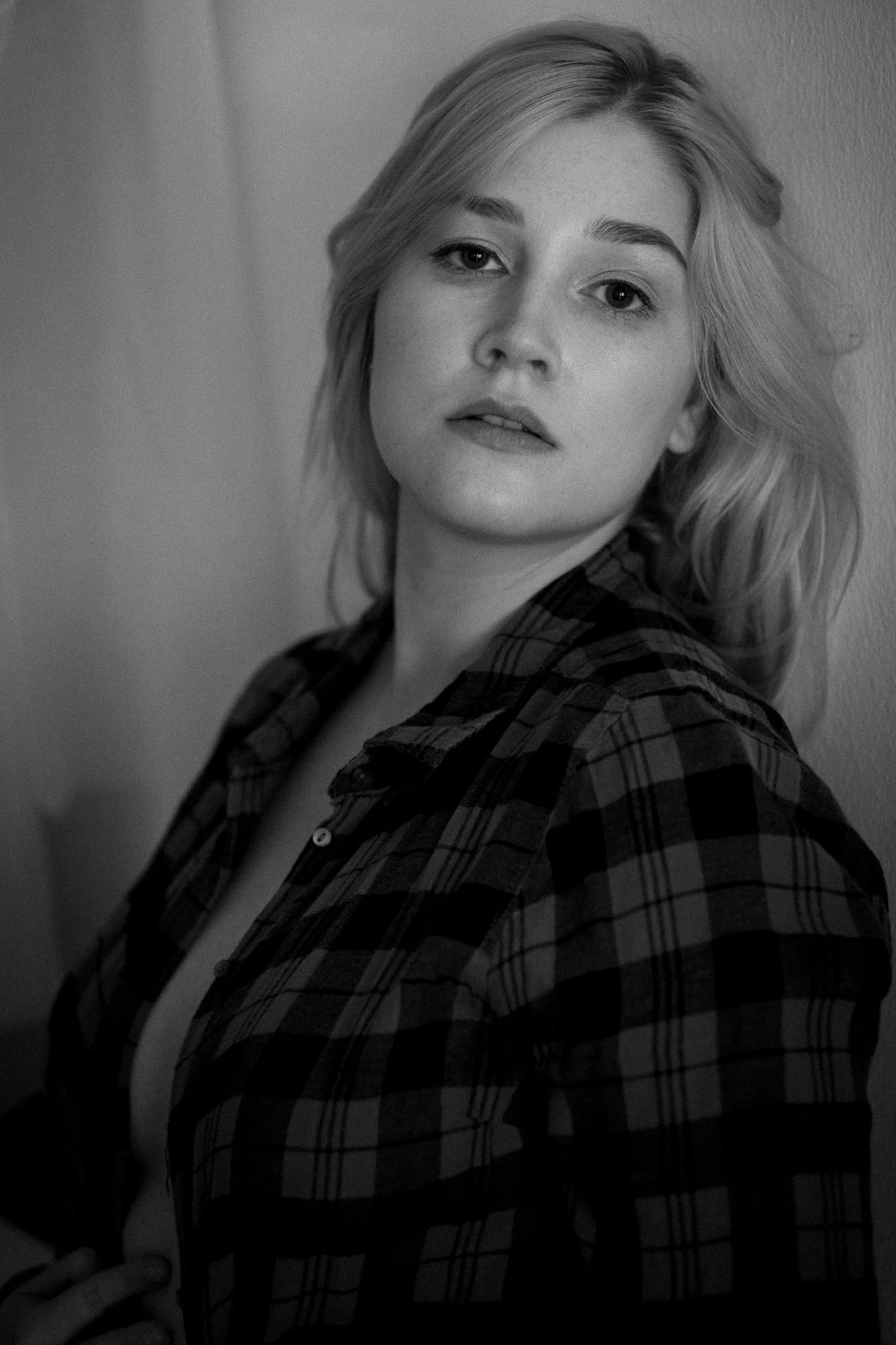 Schwarz-Weiß-Fotografie einer jungen Frau mit blonden, lockigen Haaren, die eine karierte Jacke trägt, vor einer glatten Wand.