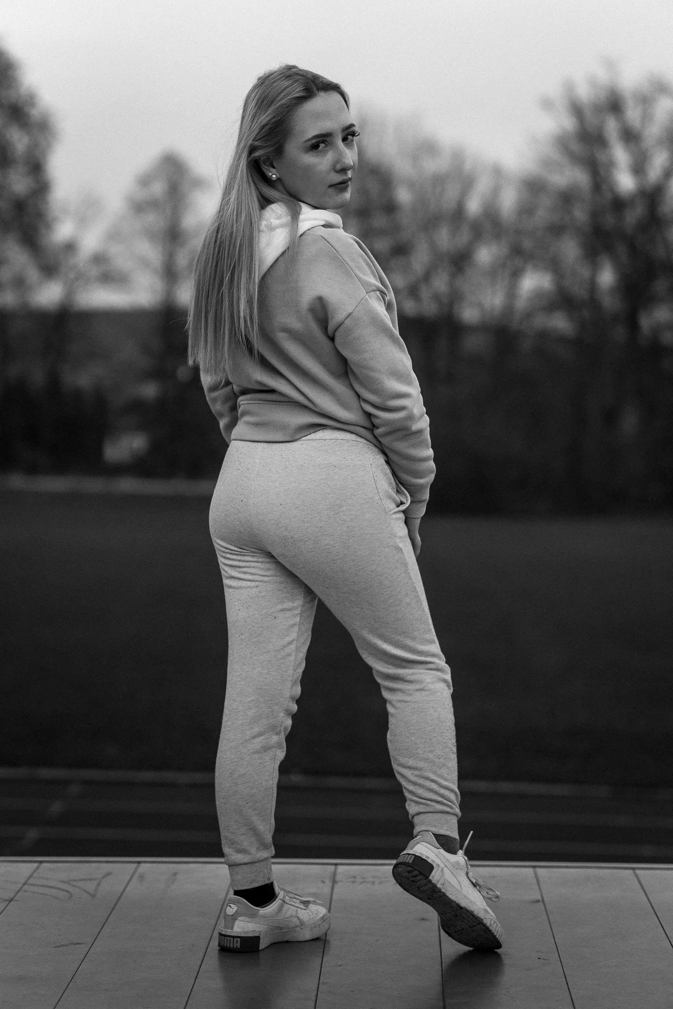 Schwarz-Weiß-Foto einer jungen Frau, die auf einem Laufband steht, trägt Sportschuhe und eine Trainingshose, mit Blick in die Kamera, im Freien, im Hintergrund Bäume.