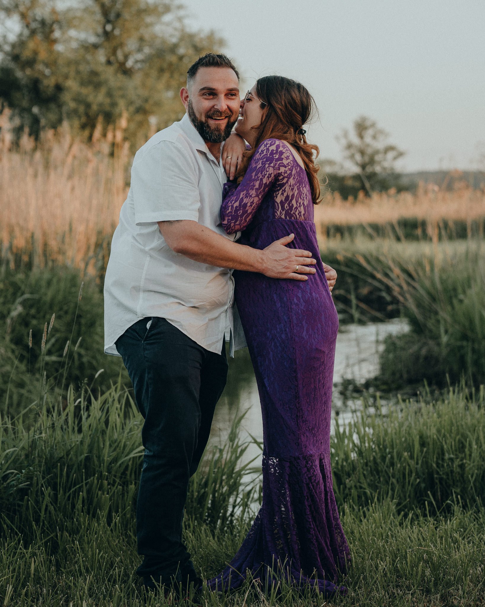 Ein lächelndes Paar umarmt sich in einer natürlichen Landschaft bei Sonnenuntergang, im Hintergrund Bäume, Gräser und ein kleiner Wasserlauf.