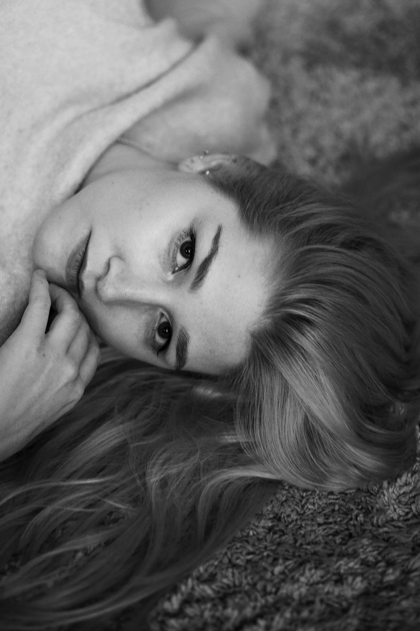 Ein Porträtfoto einer jungen Frau mit langen Haaren, die auf dem Boden liegt, mit einem nachdenklichen Ausdruck, in Schwarz-Weiß aufgenommen.