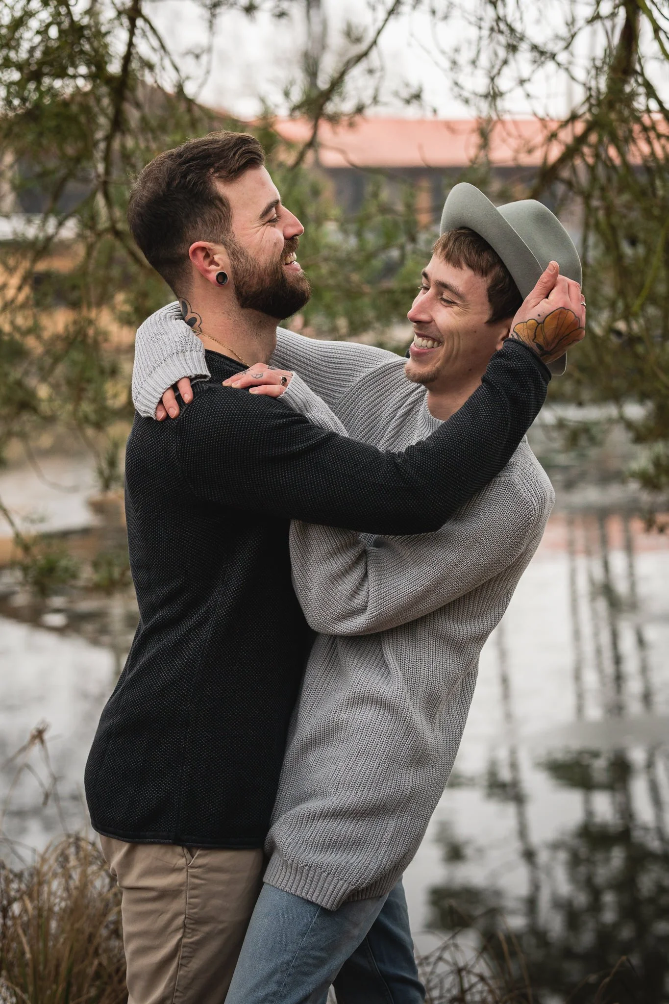 Zwei Männer, die sich umarmen und lachend sich ansehen, stehen am Wasser im Freien, umgeben von Bäumen.