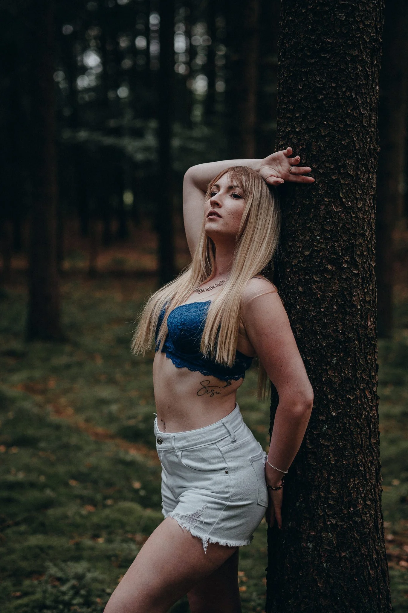 Junge Frau mit blonder Haare in einem blauen Top und weißen Shorts lehnt an einem Baum im Wald.