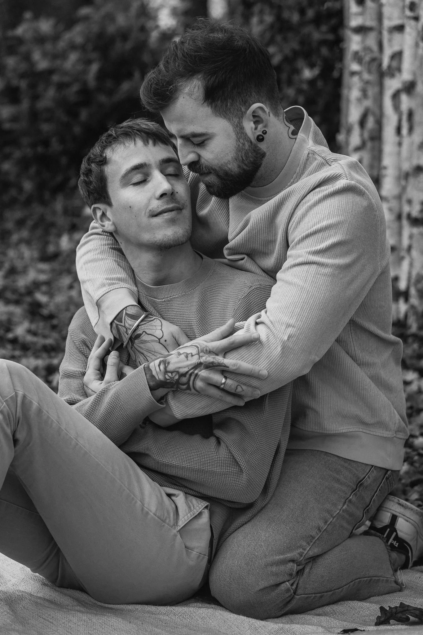 Zwei Männer sitzen im Freien, der eine hält den anderen liebevoll im Arm und beide haben die Augen geschlossen. Es ist eine intime, liebevolle Szene in Schwarzweiß.