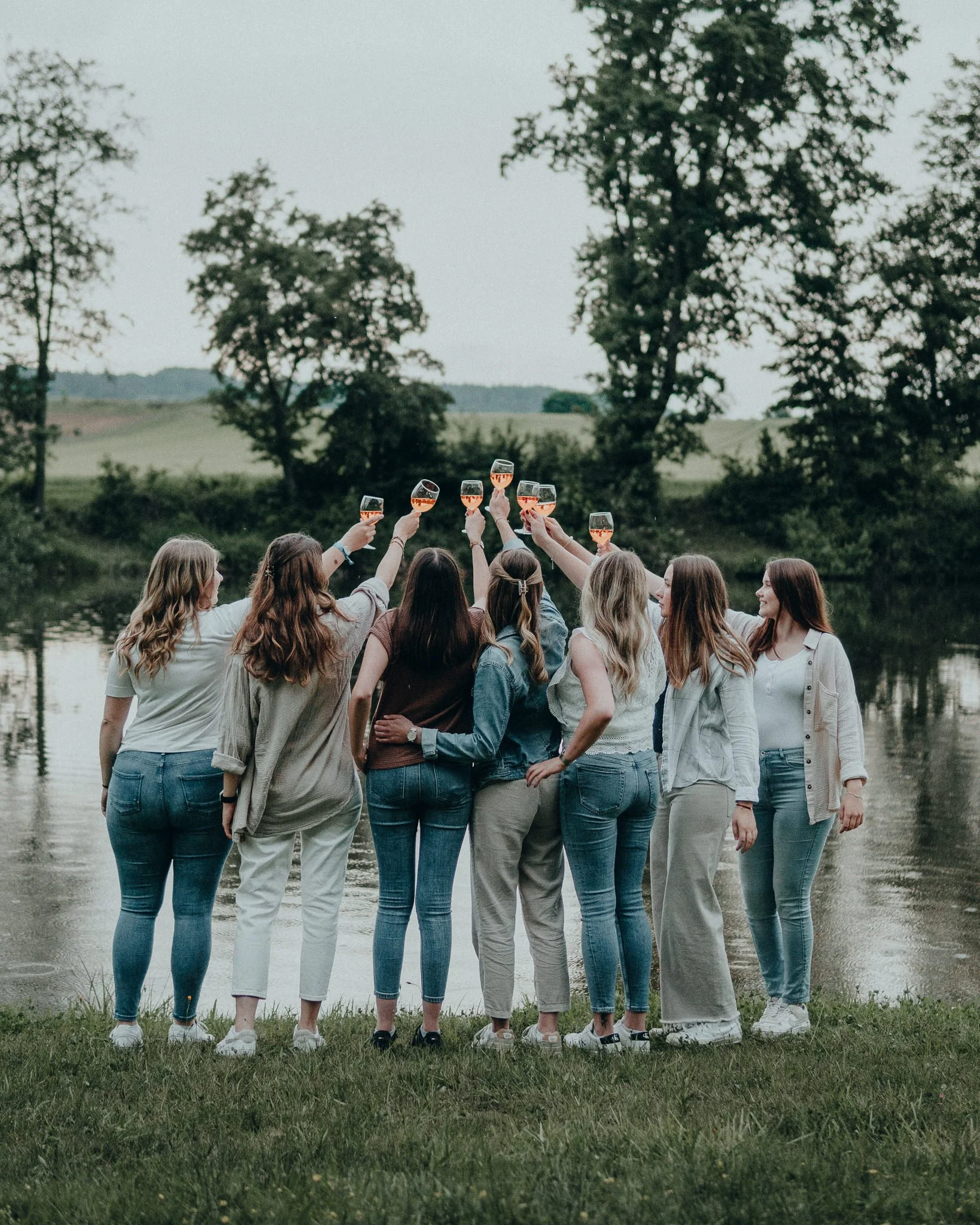 Eine Gruppe von Frauen steht am Ufer eines Flusses, hebt Gläser mit Roséwein zum Anstoßen, bei Sonnenuntergang im Freien, umgeben von Bäumen.