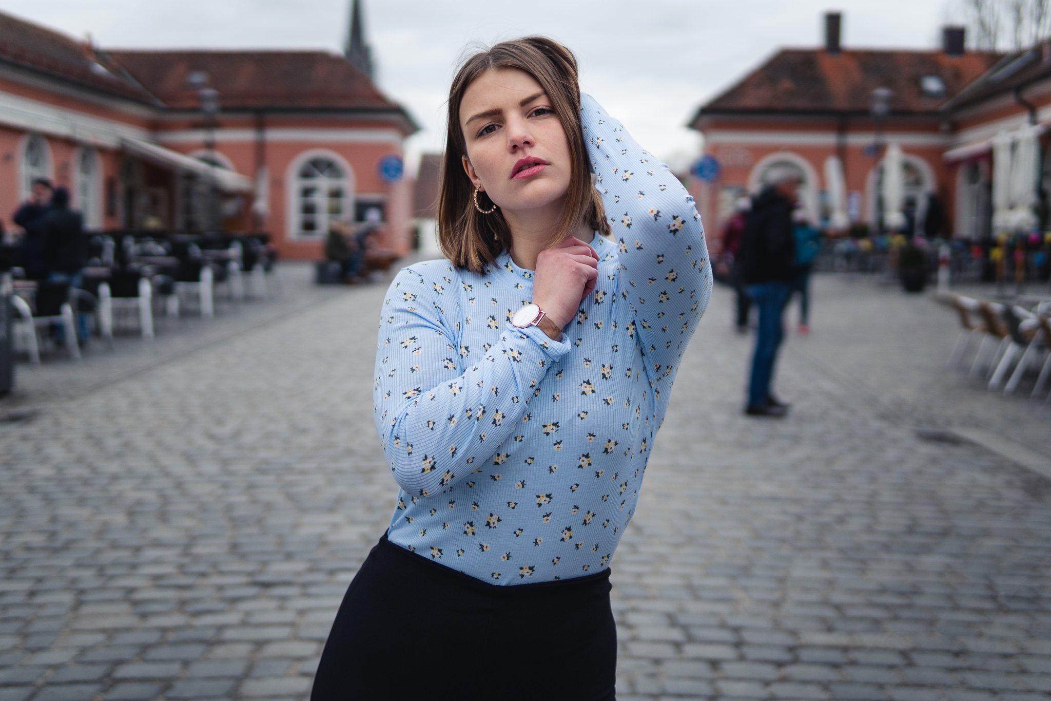Junge Frau steht auf Kopfsteinpflasterstraße in einer belebten Stadt, trägt eine hellblaue Bluse mit Blumenmuster und eine schwarze Hose, blickt nach vorne, das Haar ist schulterlang, sie trägt Ohrringe und eine Armbanduhr.