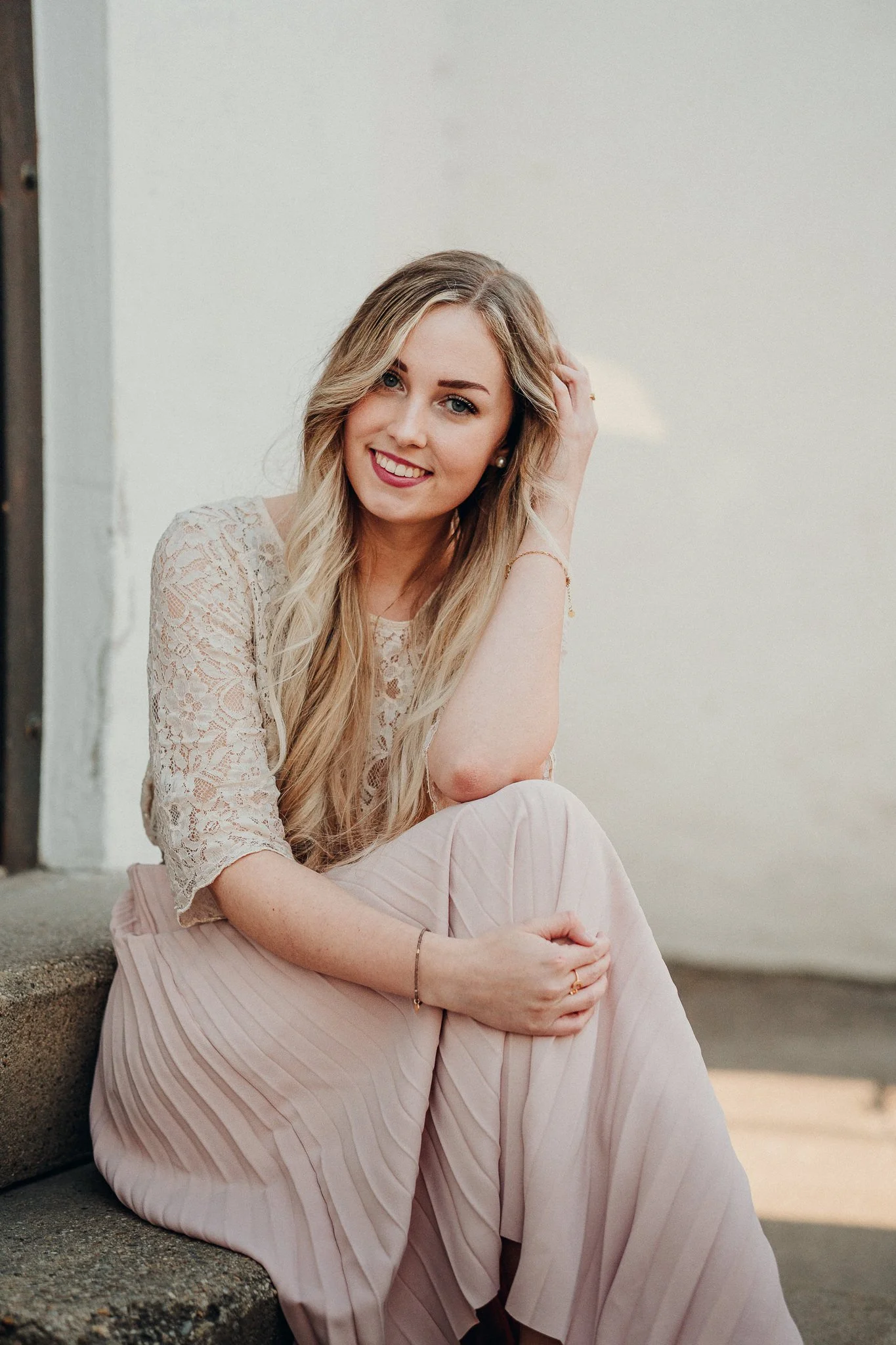 Junge Frau mit langem blonden Haar, lächelnd, im weißen Spitzenoberteil und rosa plissiertem Rock, sitzt auf einer Treppe, lehnt sich an eine Wand.